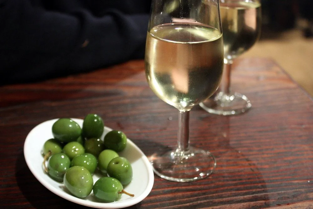 Two glasses of white wine and a small white dish with green olives on a wooden table.
