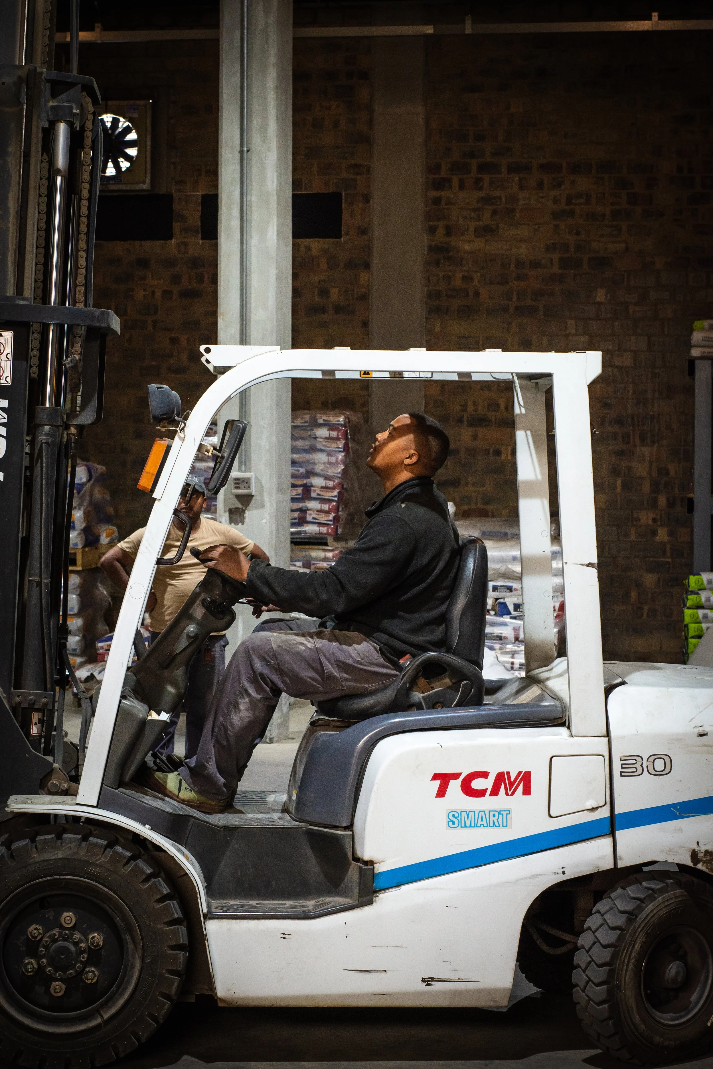 Man operating a forklift in a warehouse, wearing a black jacket and gray pants. The forklift is white with blue and red markings, and the background shows stacked bags and a brick wall.