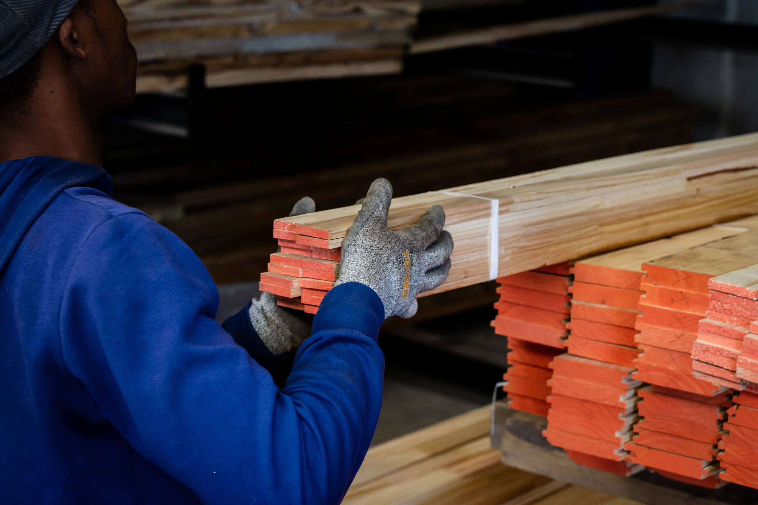 Person stacking wooden planks with painted ends in a workshop, wearing blue jacket and gloves.