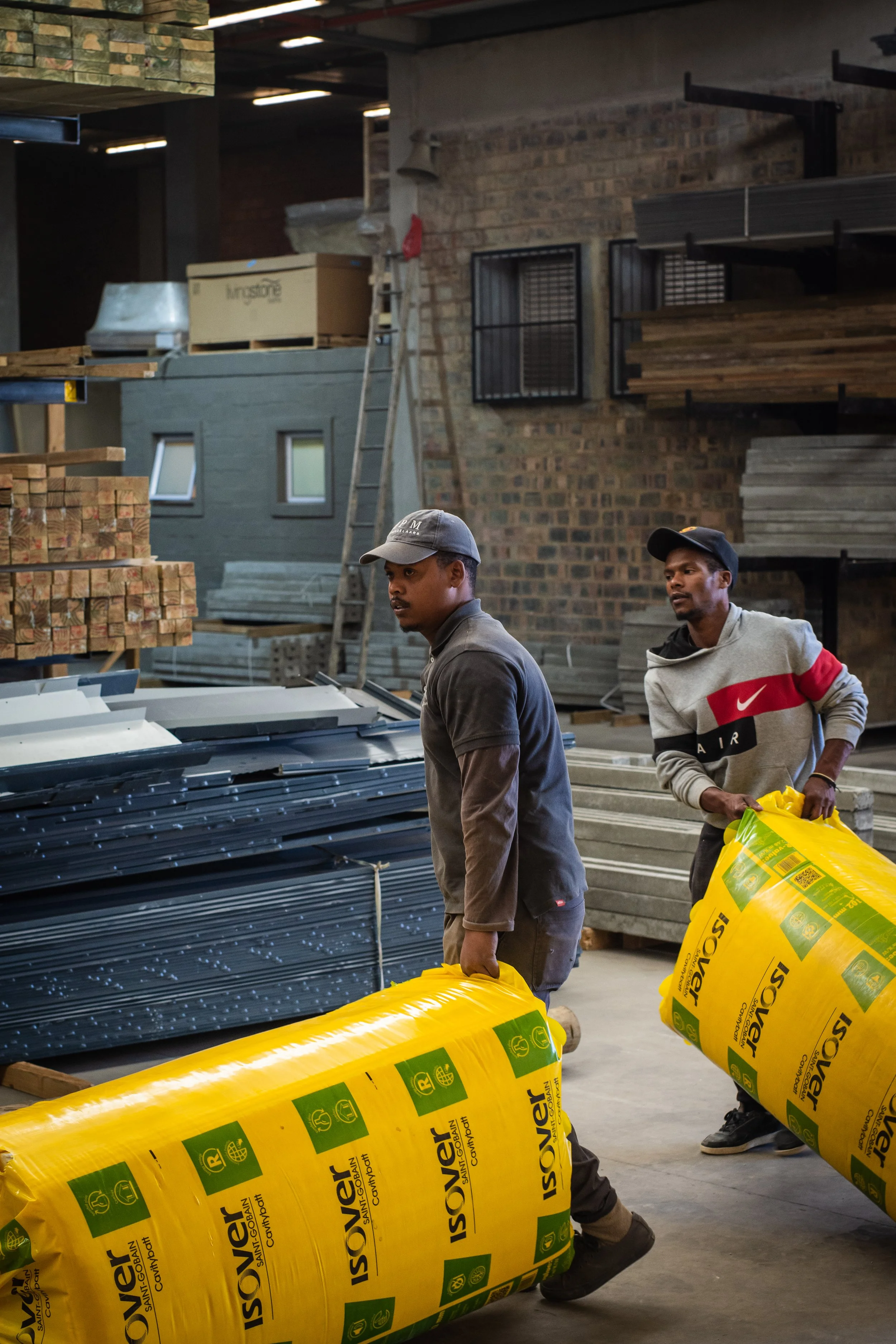 Two men carrying insulation rolls in a warehouse with stacked building materials.
