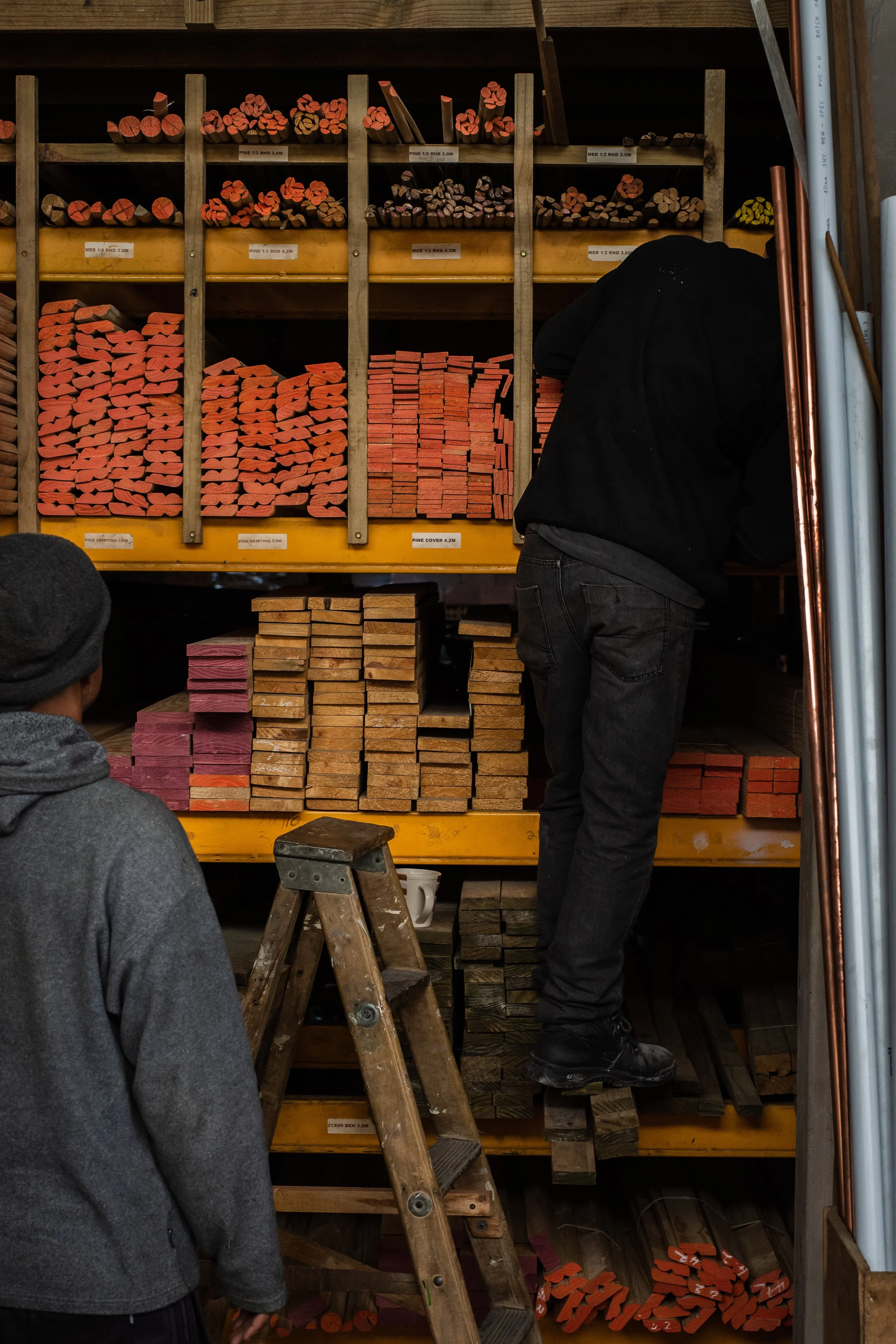 Two people organizing colorful wooden pieces on shelves in a workshop, with a wooden ladder leaning against a shelf.