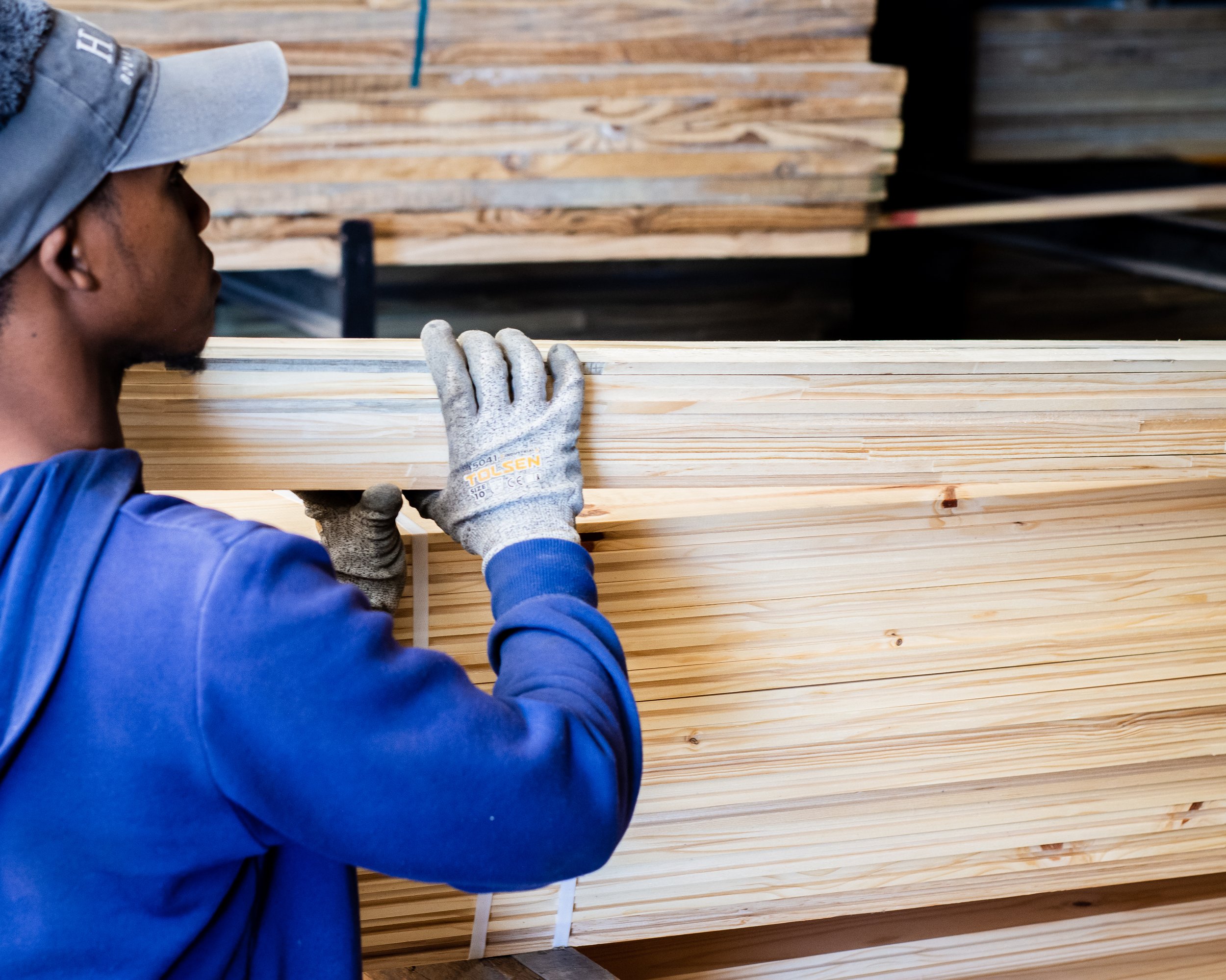 A person in a blue hoodie and cap handling a stack of wooden boards in a workshop, wearing gray gloves.