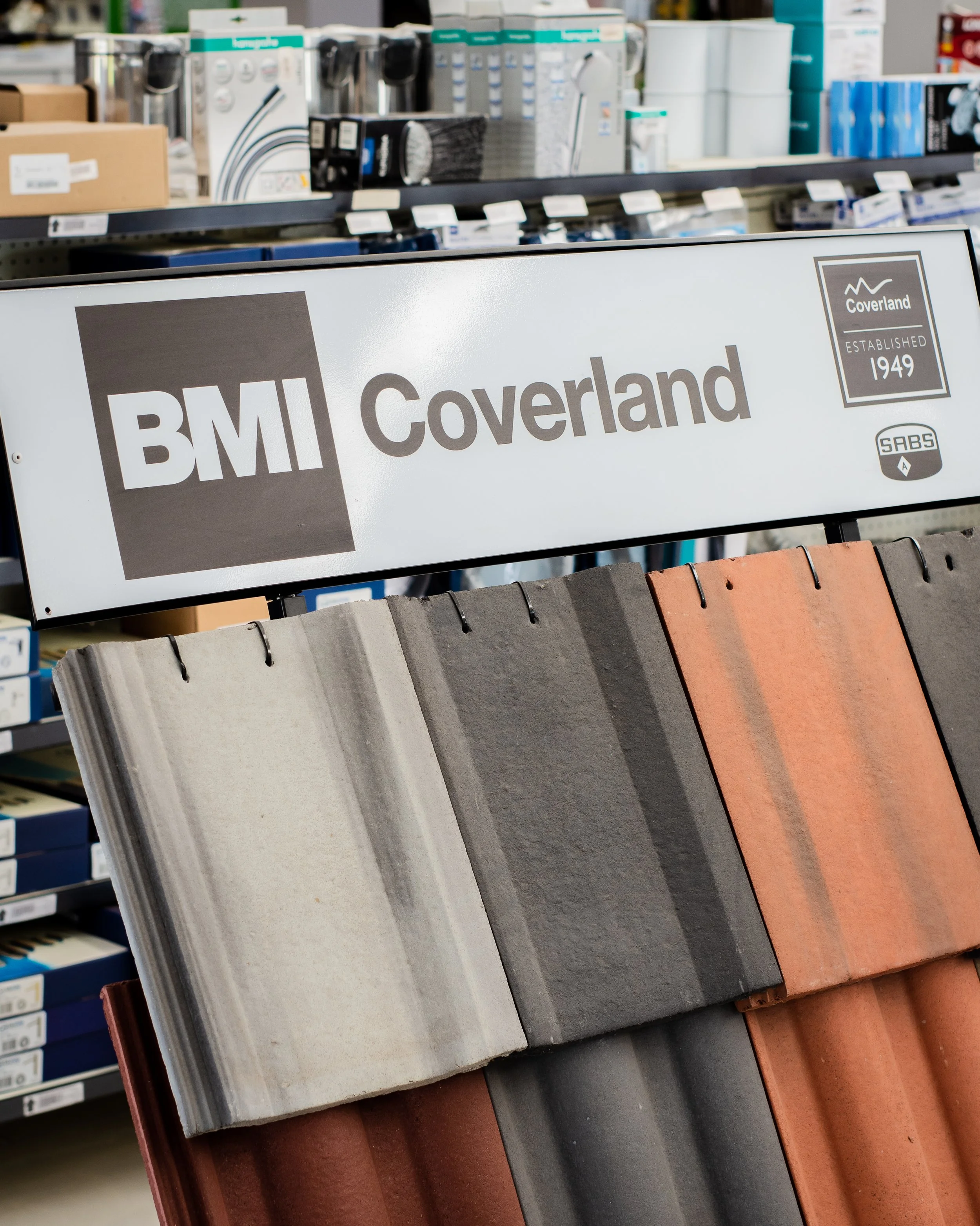 Display of roofing tiles by BMI Coverland in a store, featuring various colors and textures, with a sign indicating "BMI Coverland Established 1949."