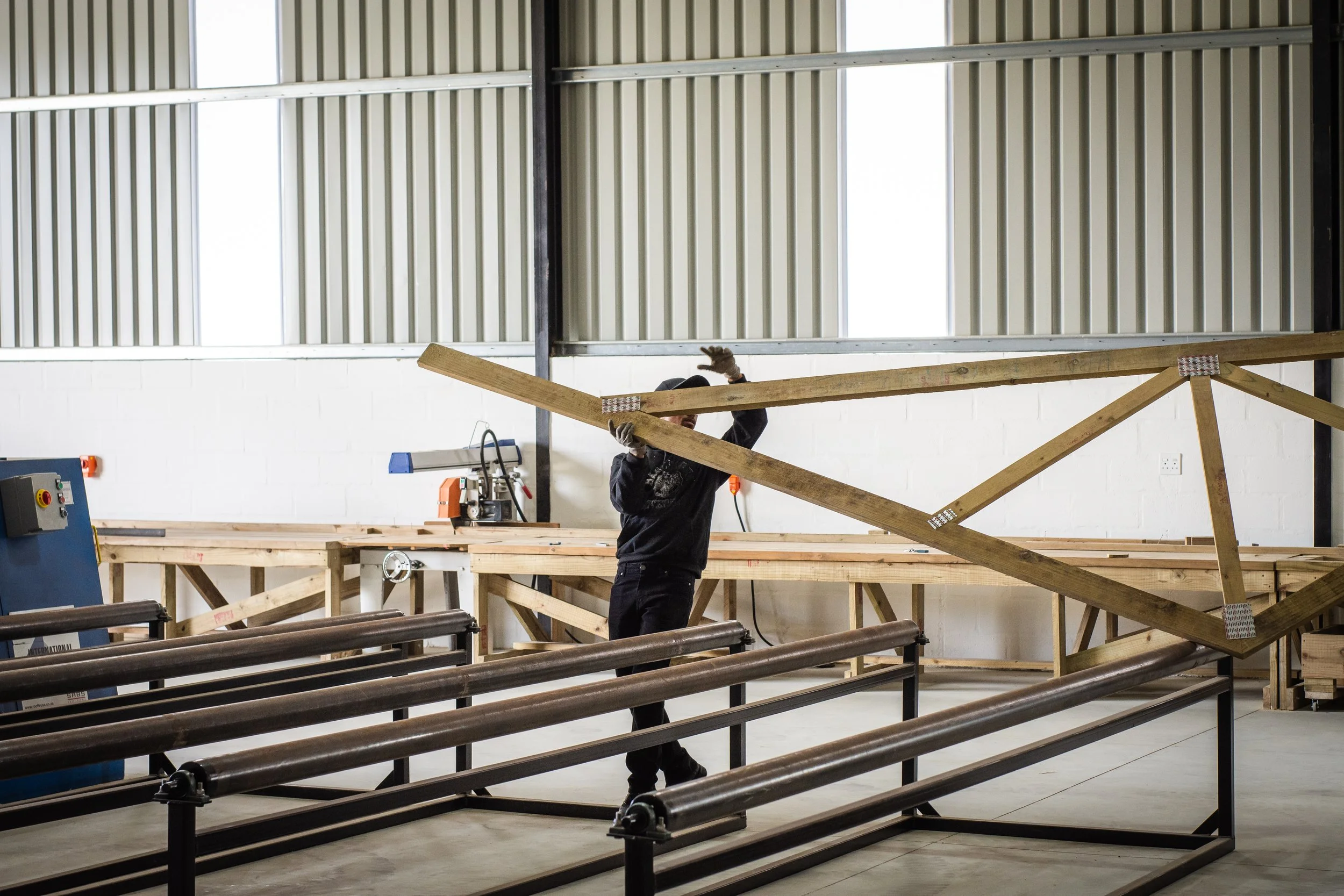 A worker in a workshop handling a wooden roof truss with metal plates, surrounded by metal supports and machinery.