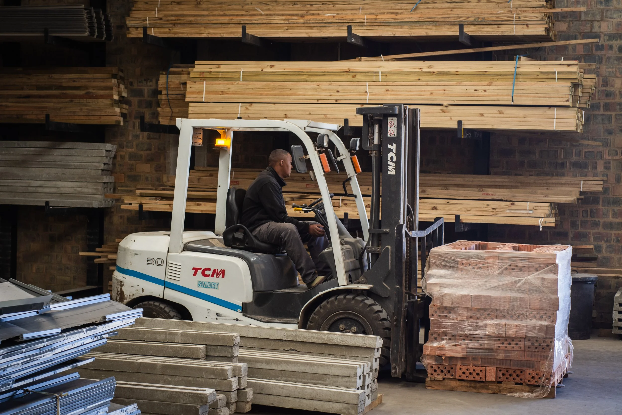 A person operating a forklift in a warehouse with stacks of wooden planks and bricks.