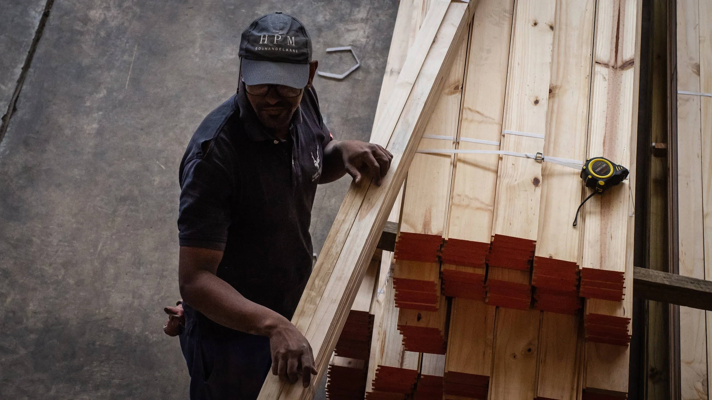 A person wearing a cap and glasses handling wooden planks, with a measuring tape visible, in a workshop or construction setting.