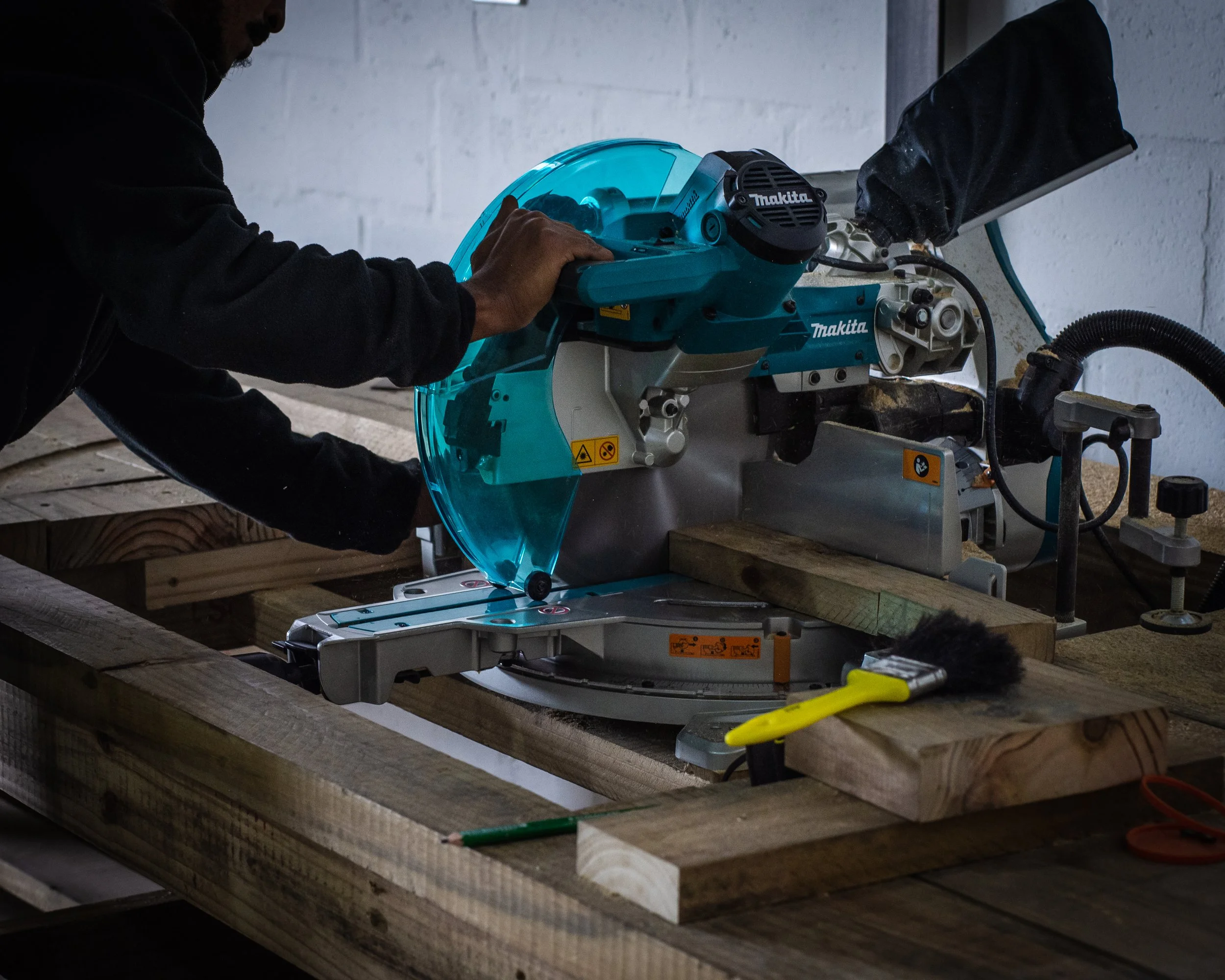 Person operating a Makita compound miter saw, cutting wood planks in a workshop.