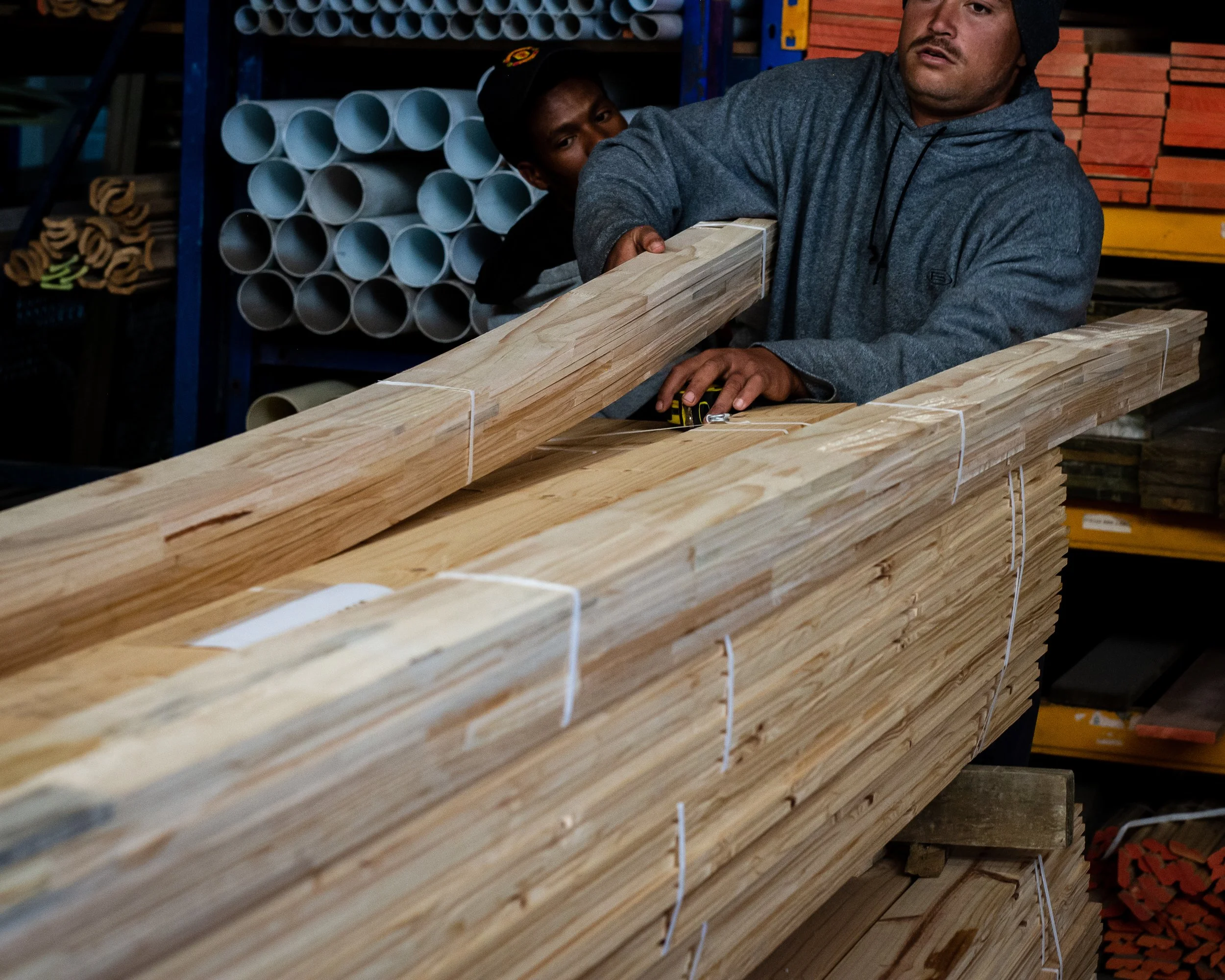 Two men handling piles of wooden planks in a warehouse environment.