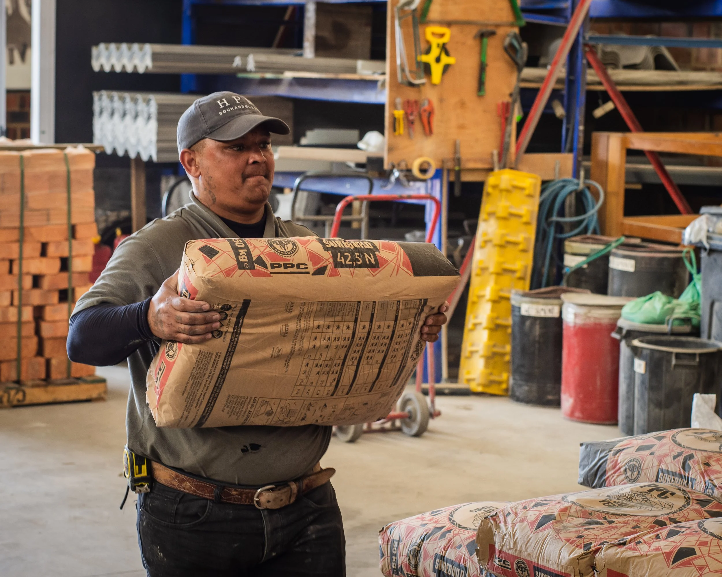 A man holding a bag of cement in a hardware store or warehouse, with tools and construction materials in the background.