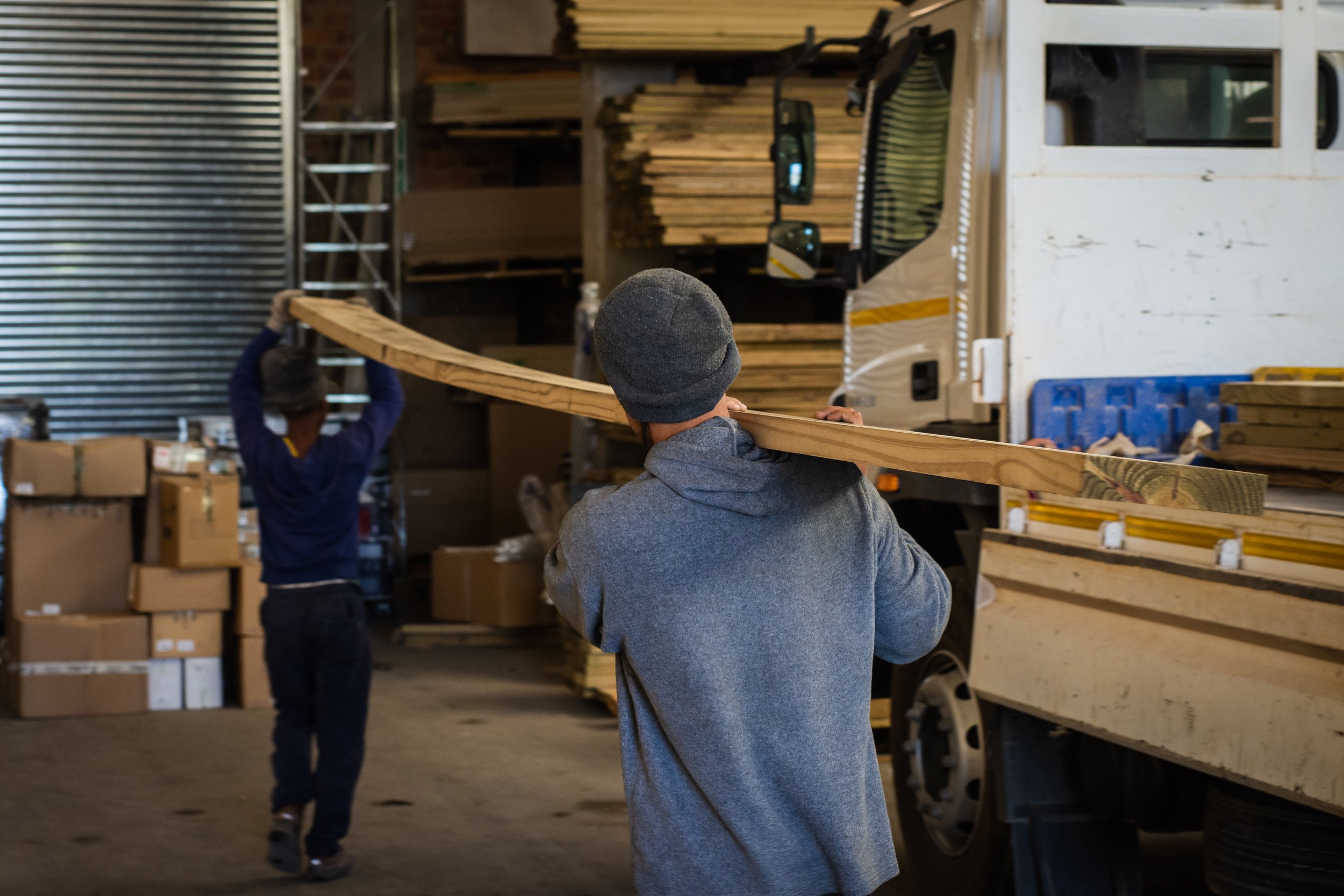 Two workers carrying a wooden plank in a warehouse with a truck and boxed inventory in the background.