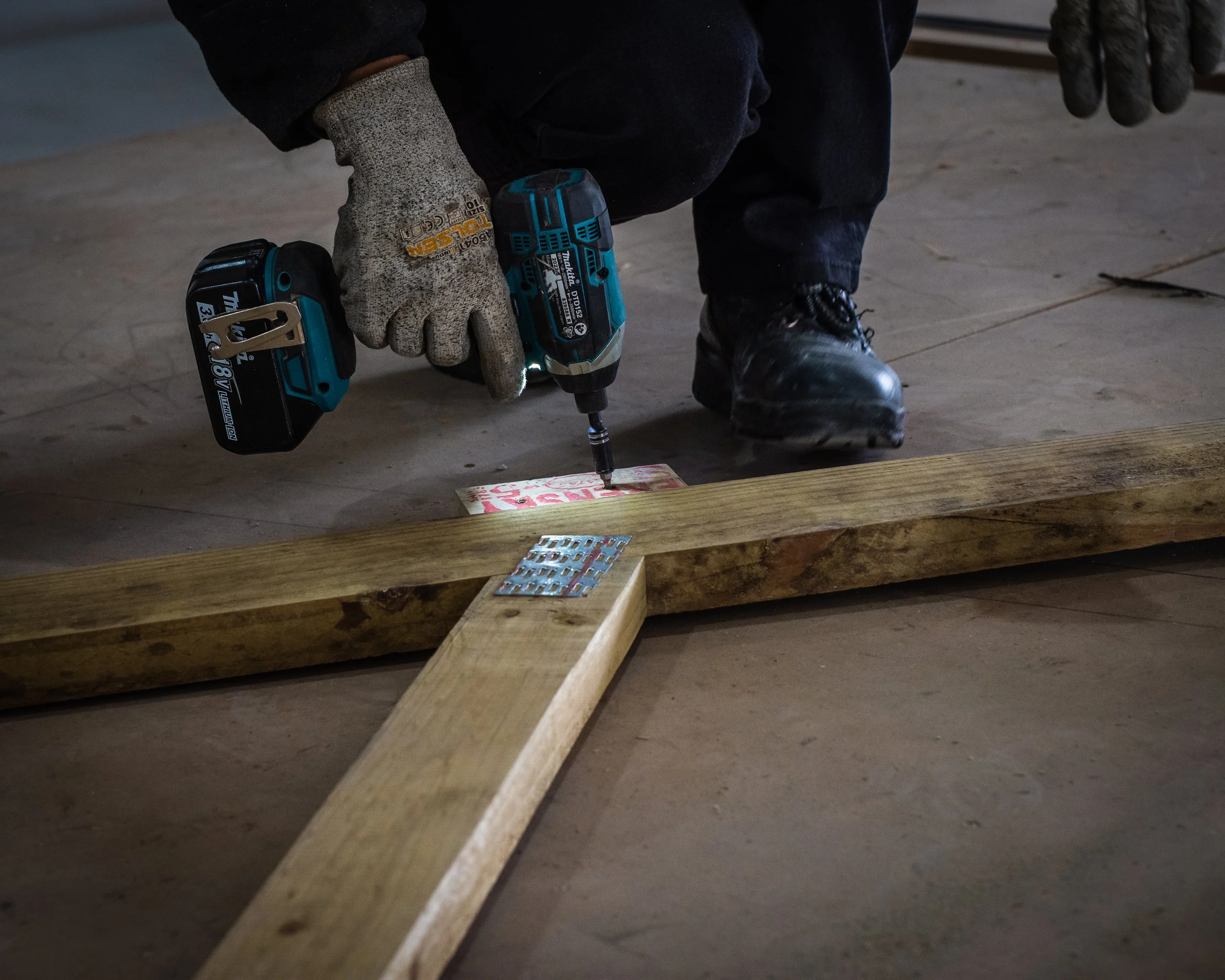 Person using a cordless drill to screw a wooden structure on the floor, wearing gloves and black boots.