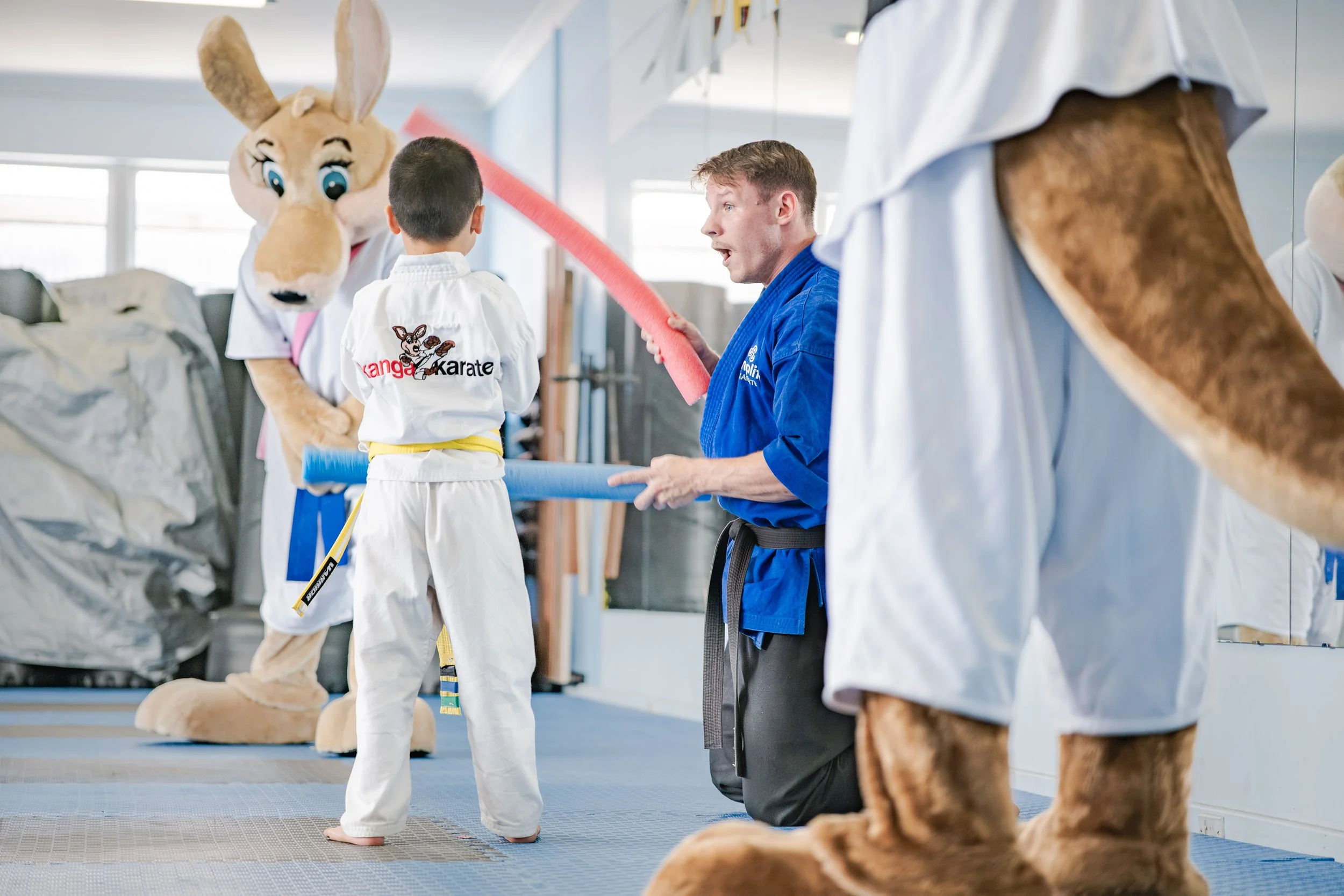 A child in a karate uniform with a yellow belt is practicing with a man in a blue karate gi who is holding a foam noodle. A person in an oversized kangaroo costume stands nearby in a martial arts dojo.