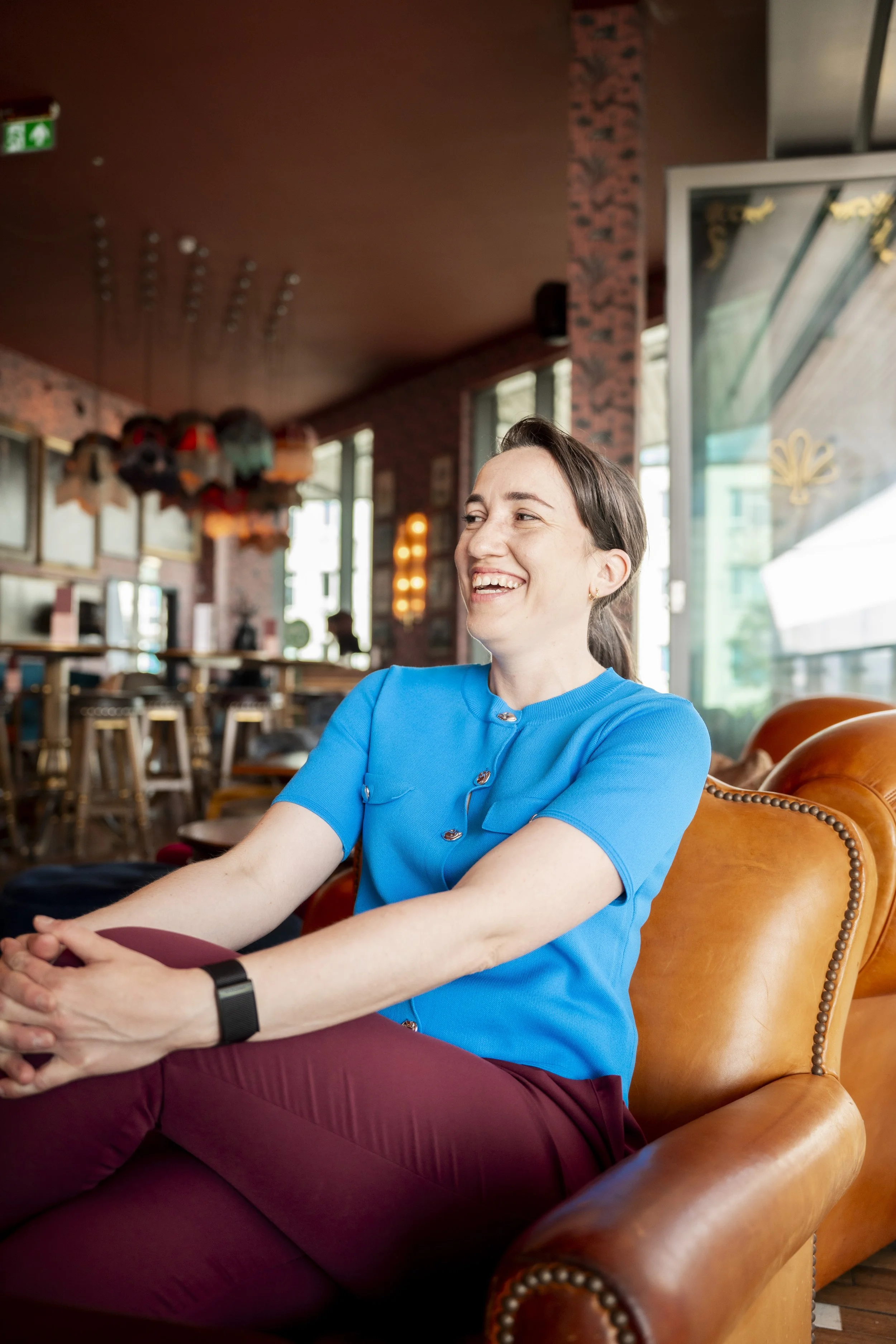 A woman with brown hair, wearing a blue top and maroon pants, smiling and sitting on a brown leather armchair in a cozy, stylish café with red walls, large windows, and decorative items hanging from the ceiling.