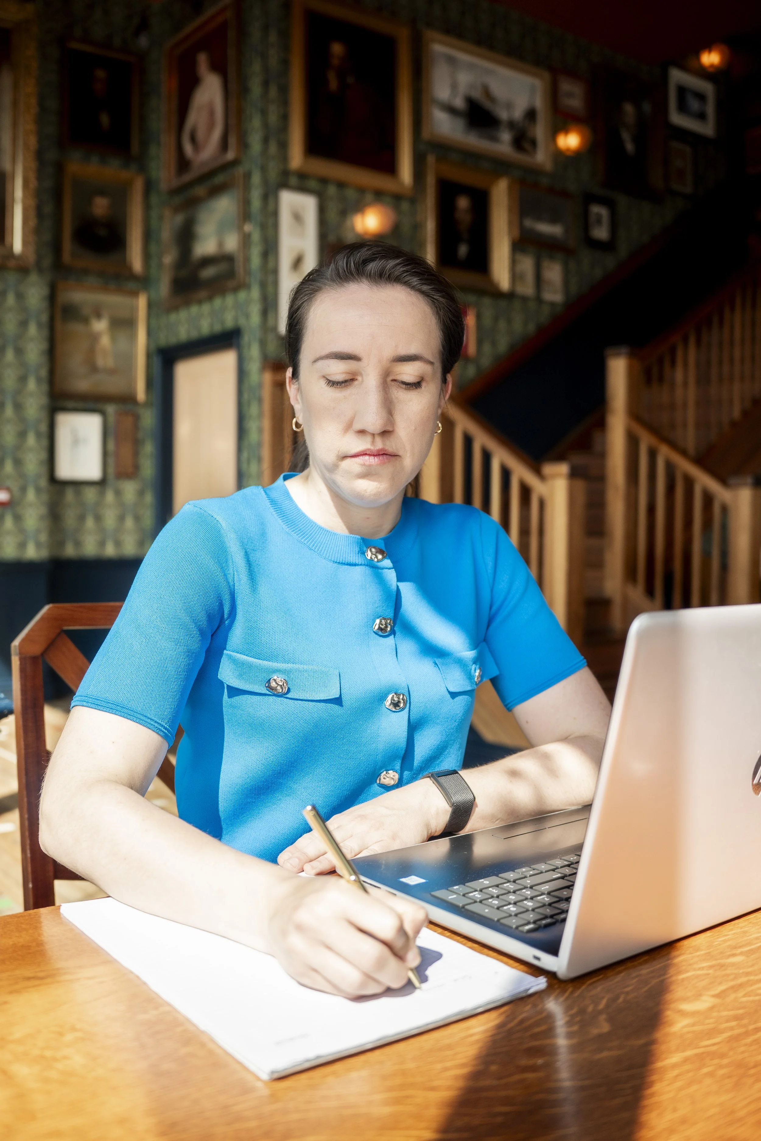 A woman in a blue top writing on a notepad with a laptop open in front of her, sitting at a wooden table in a room with framed pictures on the wall.