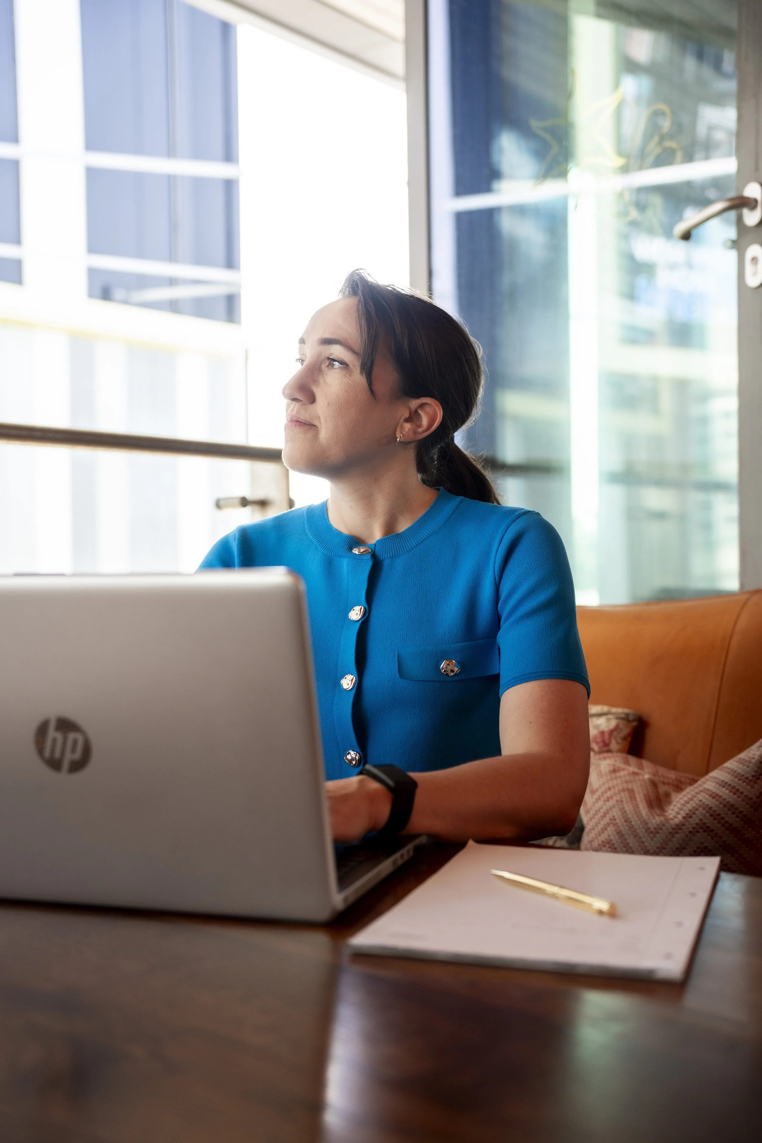 A woman with dark hair sitting at a wooden table working on a silver HP laptop in a room with large windows and modern building views.
