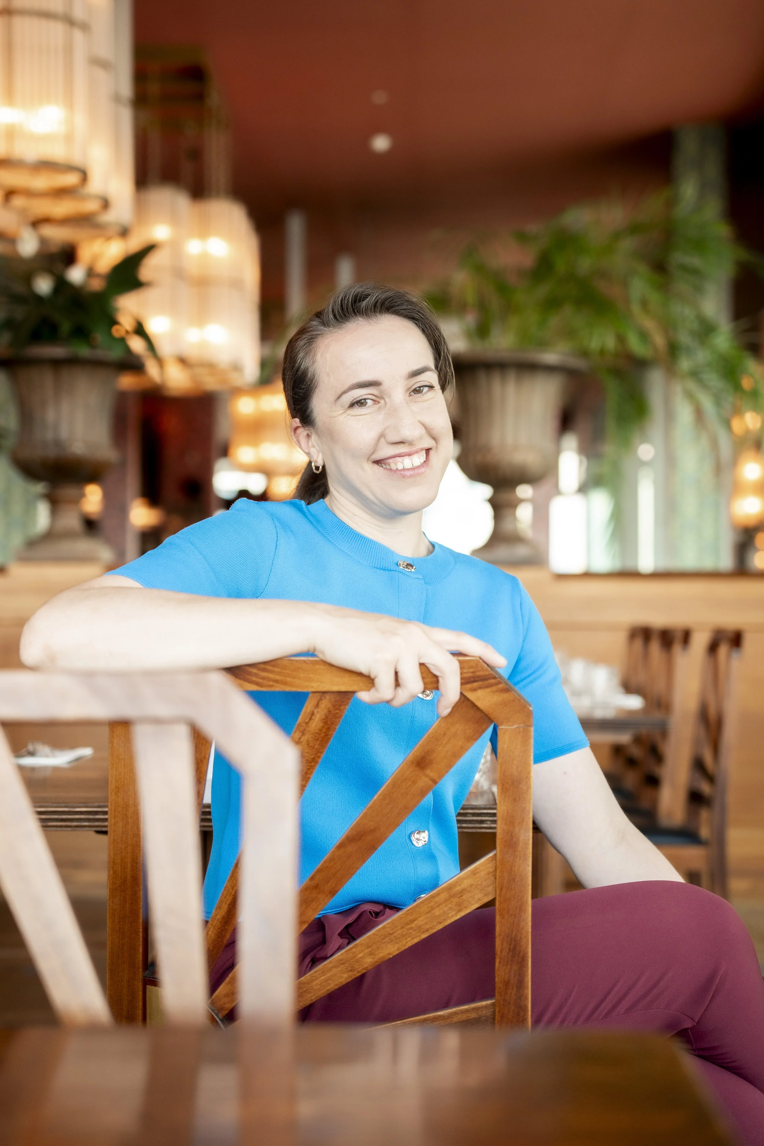 A smiling woman in a blue shirt sitting at a wooden table in a warmly lit restaurant with potted plants and wooden decor.