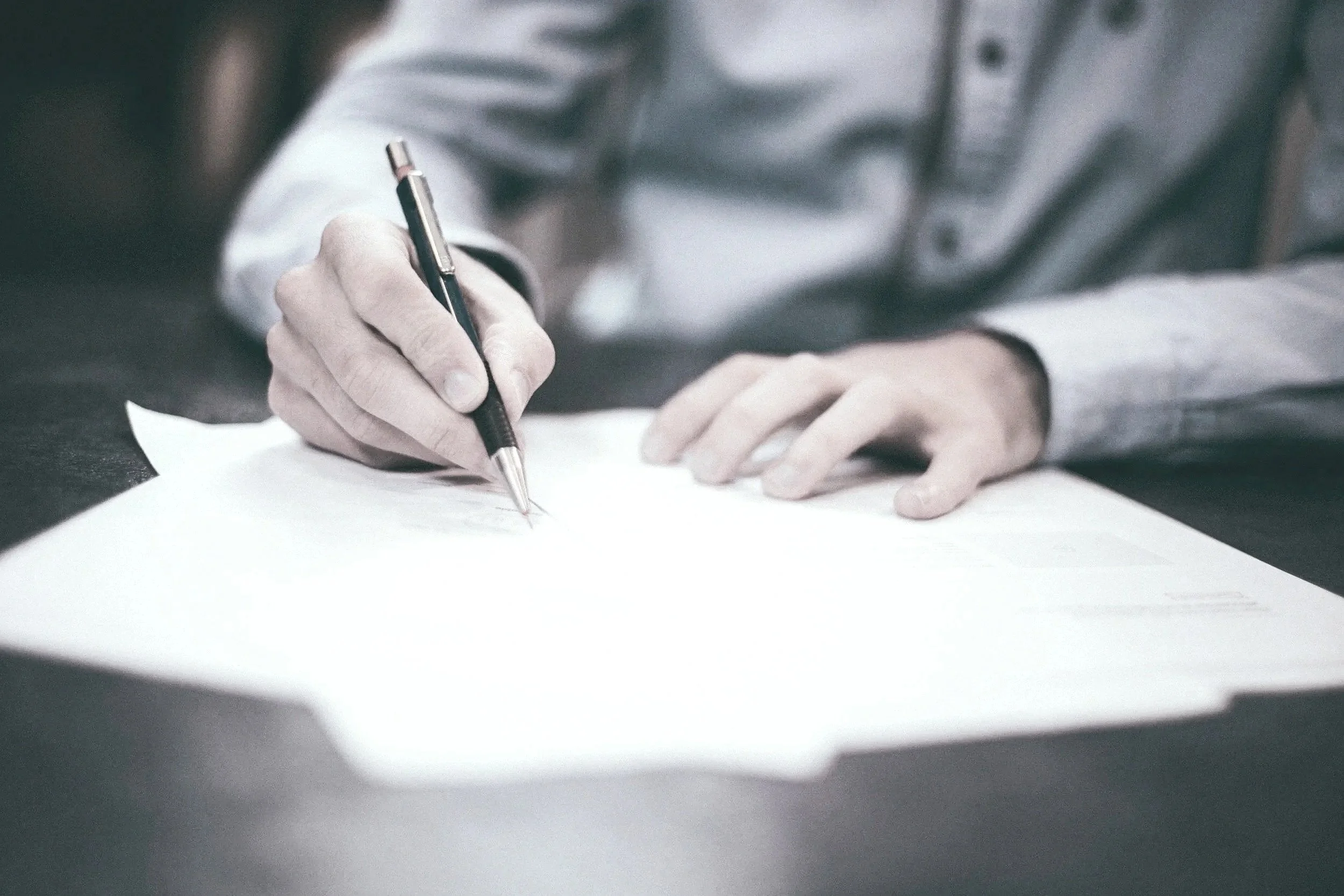 Close-up of a person wearing a light-colored shirt, writing with a black pen on a sheet of paper, with a dark surface beneath.