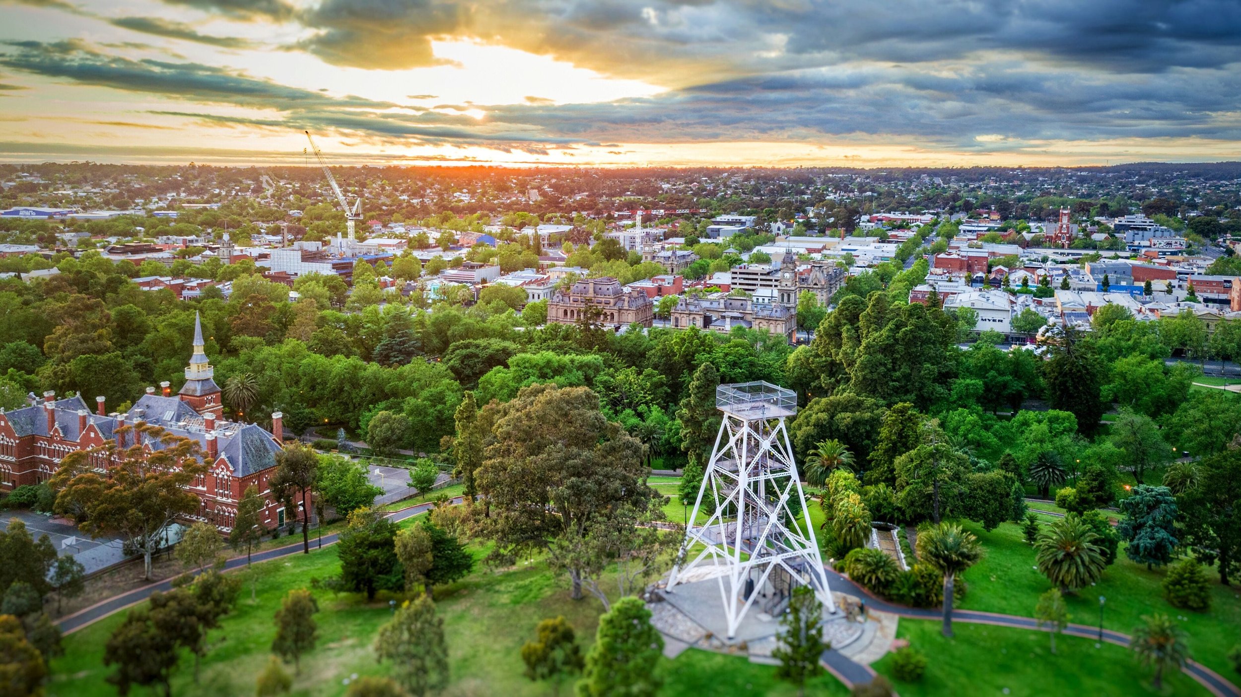 A cityscape at sunset in Cheltenham, Melbourne, with a mix of historic buildings, a park with lush green trees, and a water tower, under a partly cloudy sky.