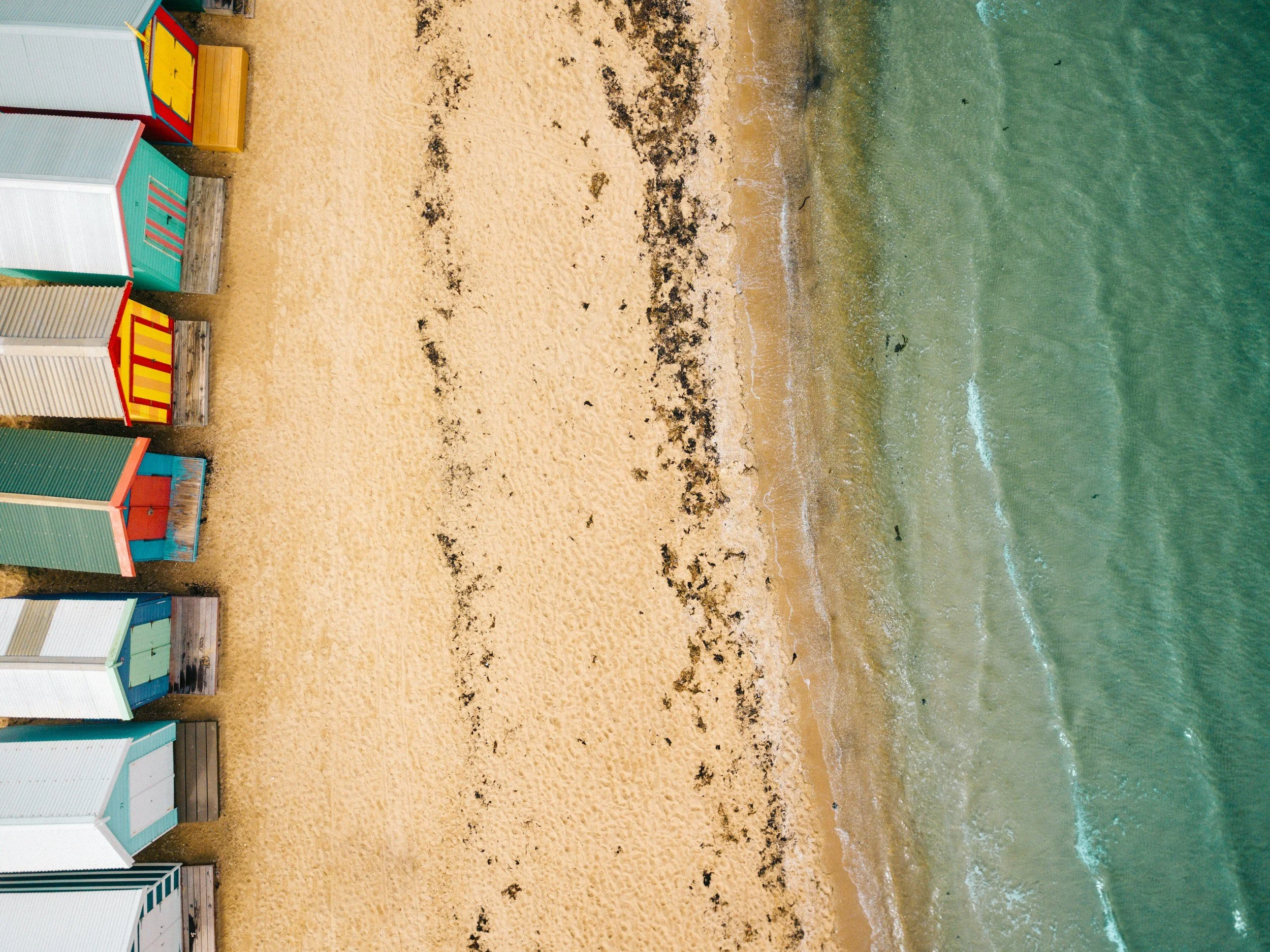 Colorful Brighton Beach bathing boxes in Melbourne lined up on the sandy shore, with seaweed and rocks scattered, and ocean waves in the background.