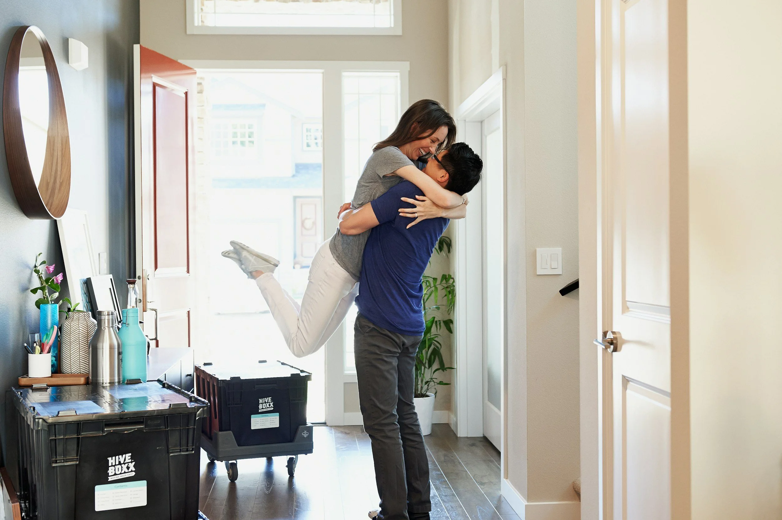 A man lifting a woman in a hug inside a bright entryway of a house, with a table with various bottles on it and moving containers in the foreground.