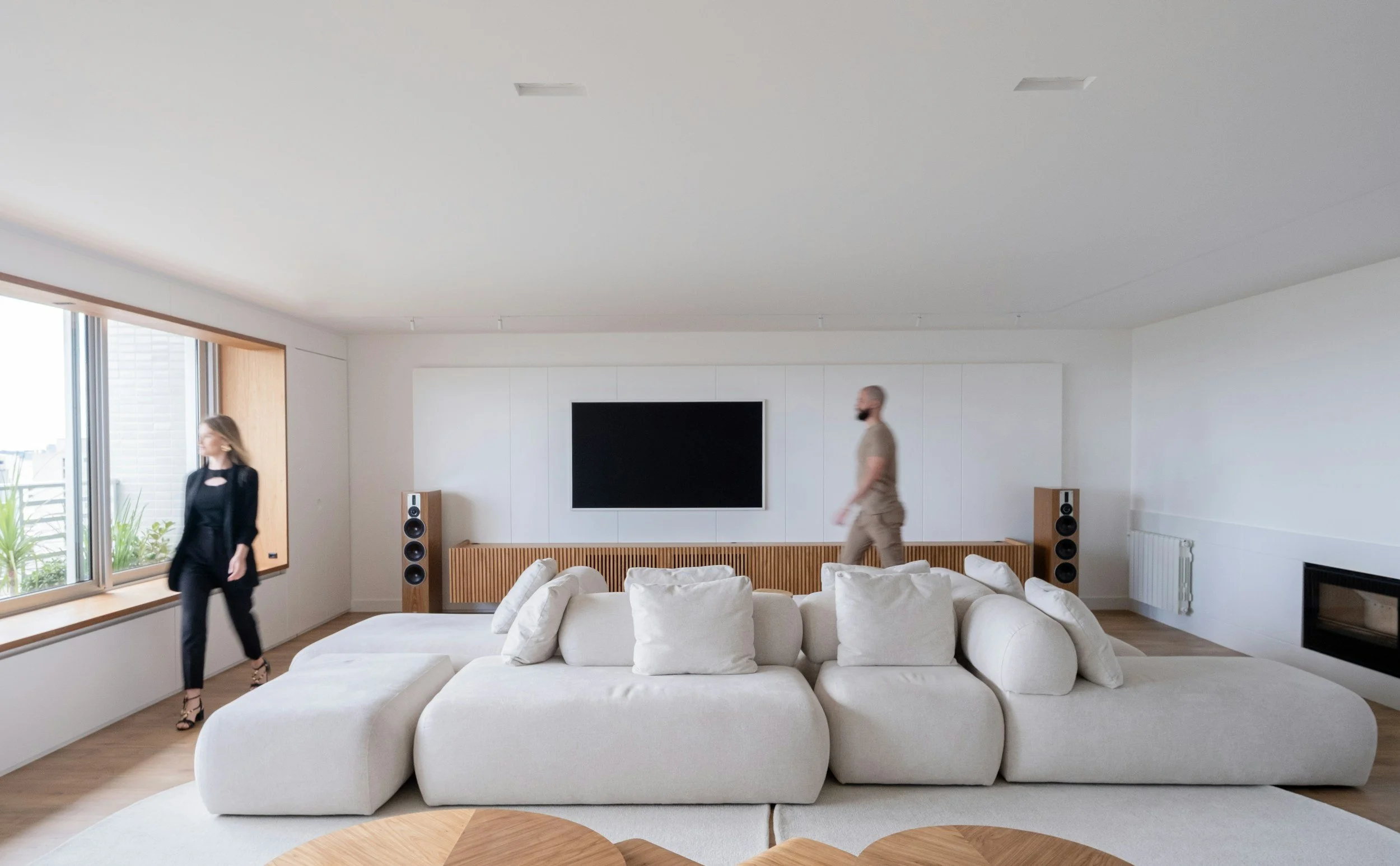 Modern living room with a large white sectional sofa, a mounted flat-screen TV, and two tall speakers. There are two people walking in the room.