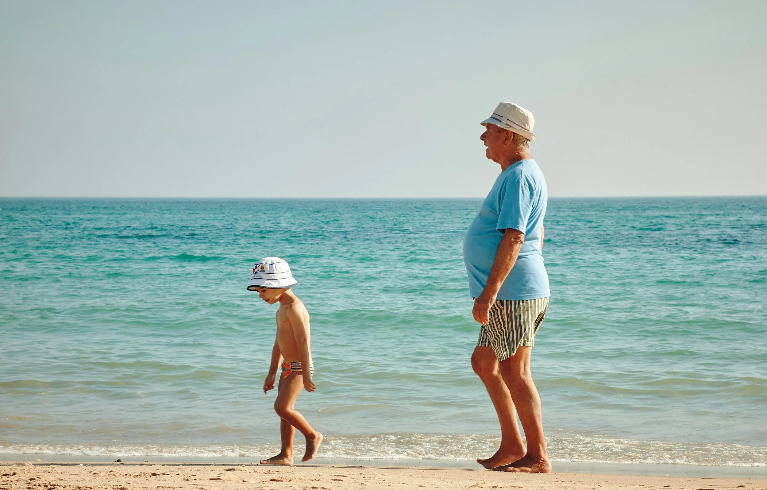 An elderly man and a young boy walking along the shoreline at the beach, both wearing hats, with the ocean and sky in the background.