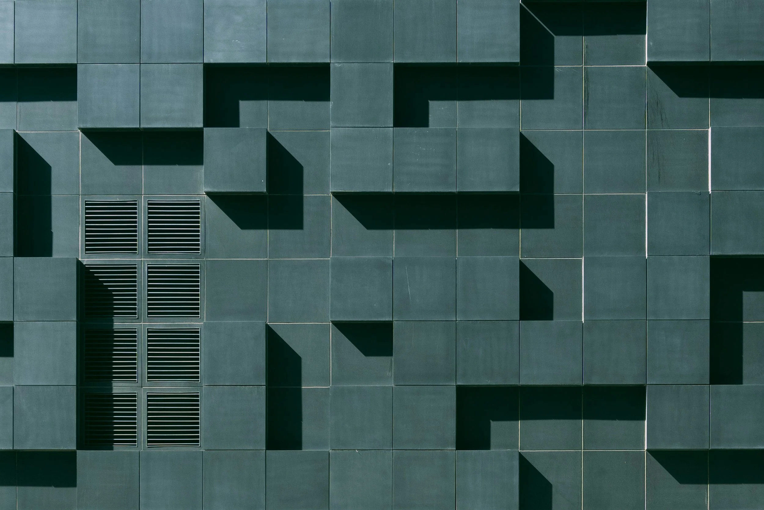 Close-up view of a modern building's green tiled wall with some tiles protruding to create a geometric pattern and vents at the bottom left.