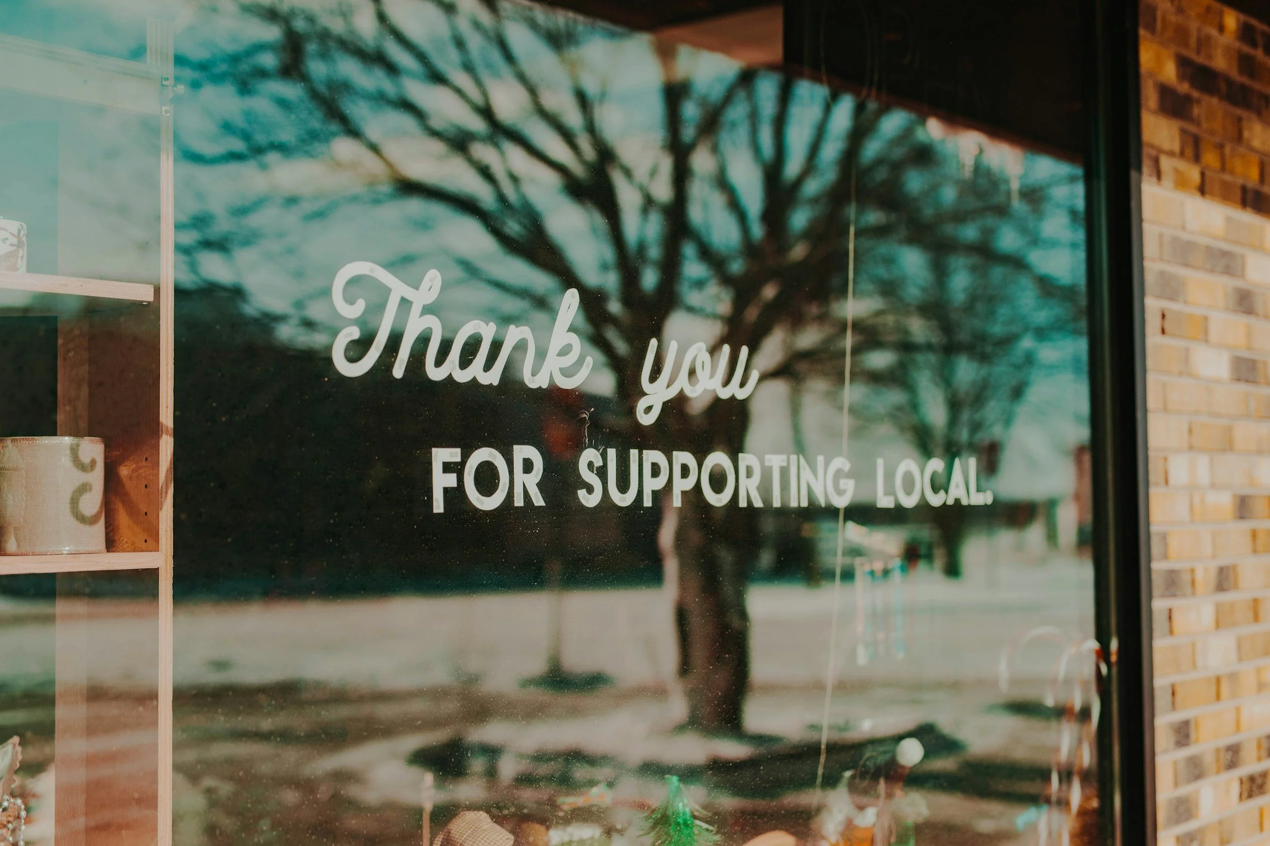 A storefront window with a reflection of a tree outside, and a sign that reads 'Thank you for supporting local.' next to a wooden shelf with a ceramic cup.