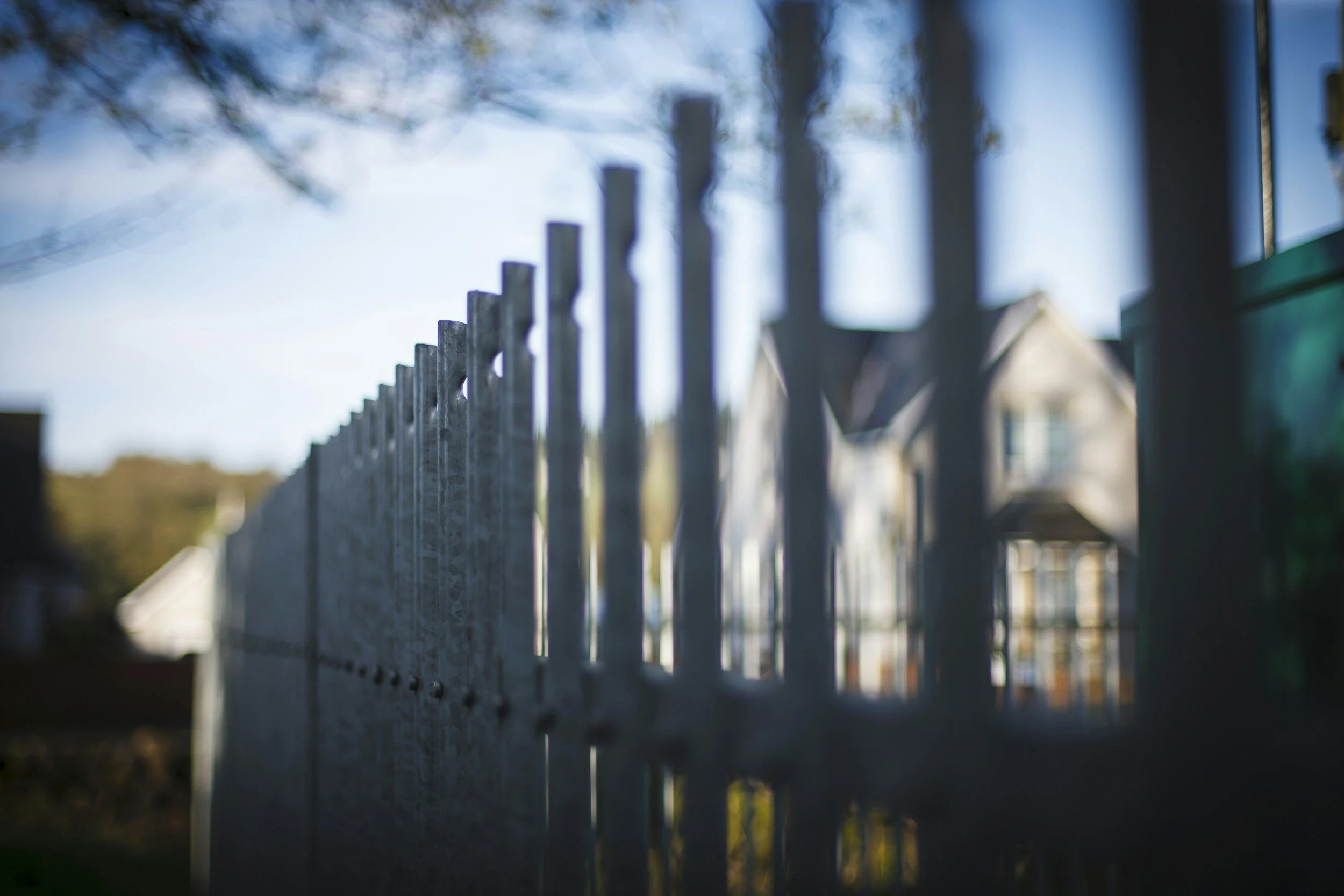 Close-up of a metal fence with pointed tops, blurred background of a house behind the fence.