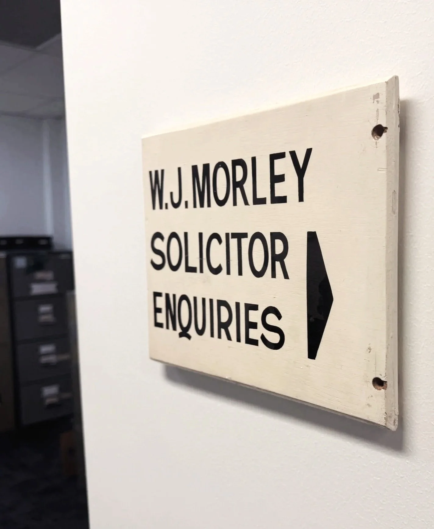 Wooden sign with black text reading 'W.J. MORLEY SOLICITOR ENQUIRIES' pointed by a black arrow, mounted on a wall.