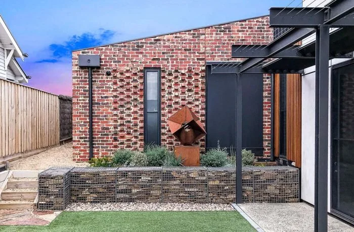 Modern backyard of a McKinnon, Melbourne home, with brick wall, small garden bed with greenery, outdoor sculpture, and a black privacy screen under a clear blue sky.