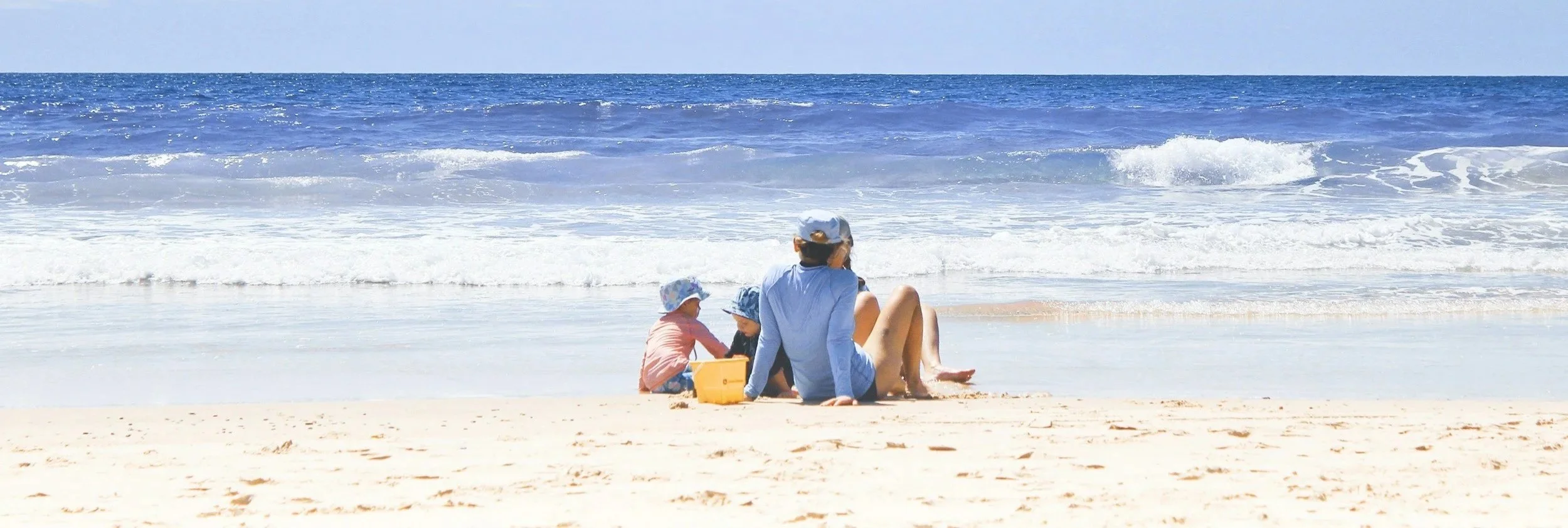 A woman and two children sitting on the sandy beach near the ocean, wearing hats, with blue water and waves in the background.