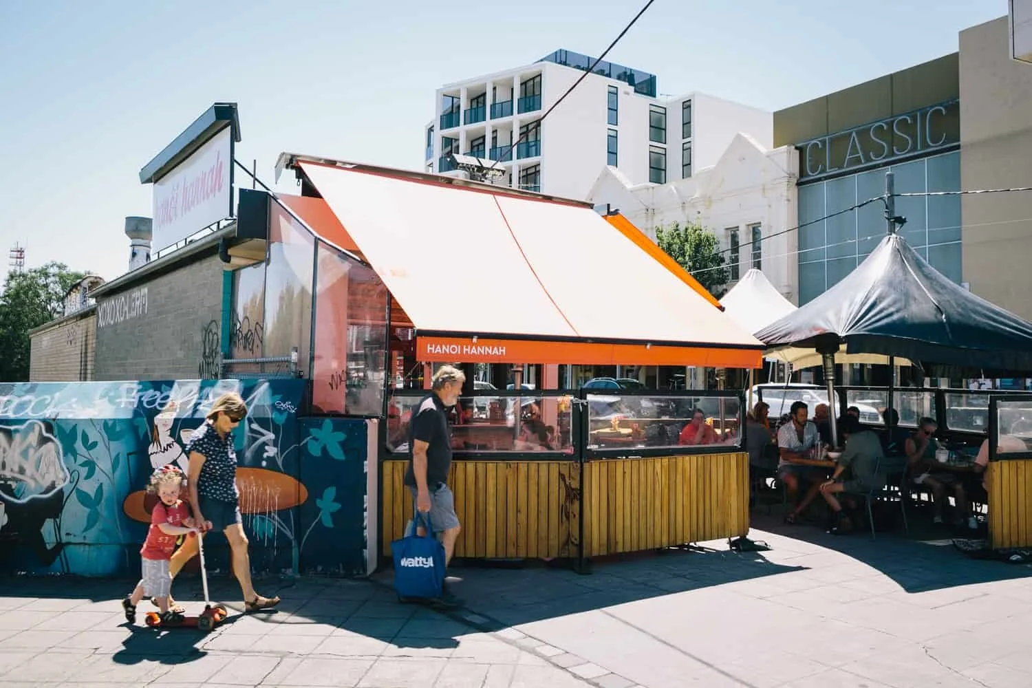 Glen Huntly Road Elsternwick, with an outdoor cafe with people dining under umbrellas, a woman and child walking past, city buildings in the background.
