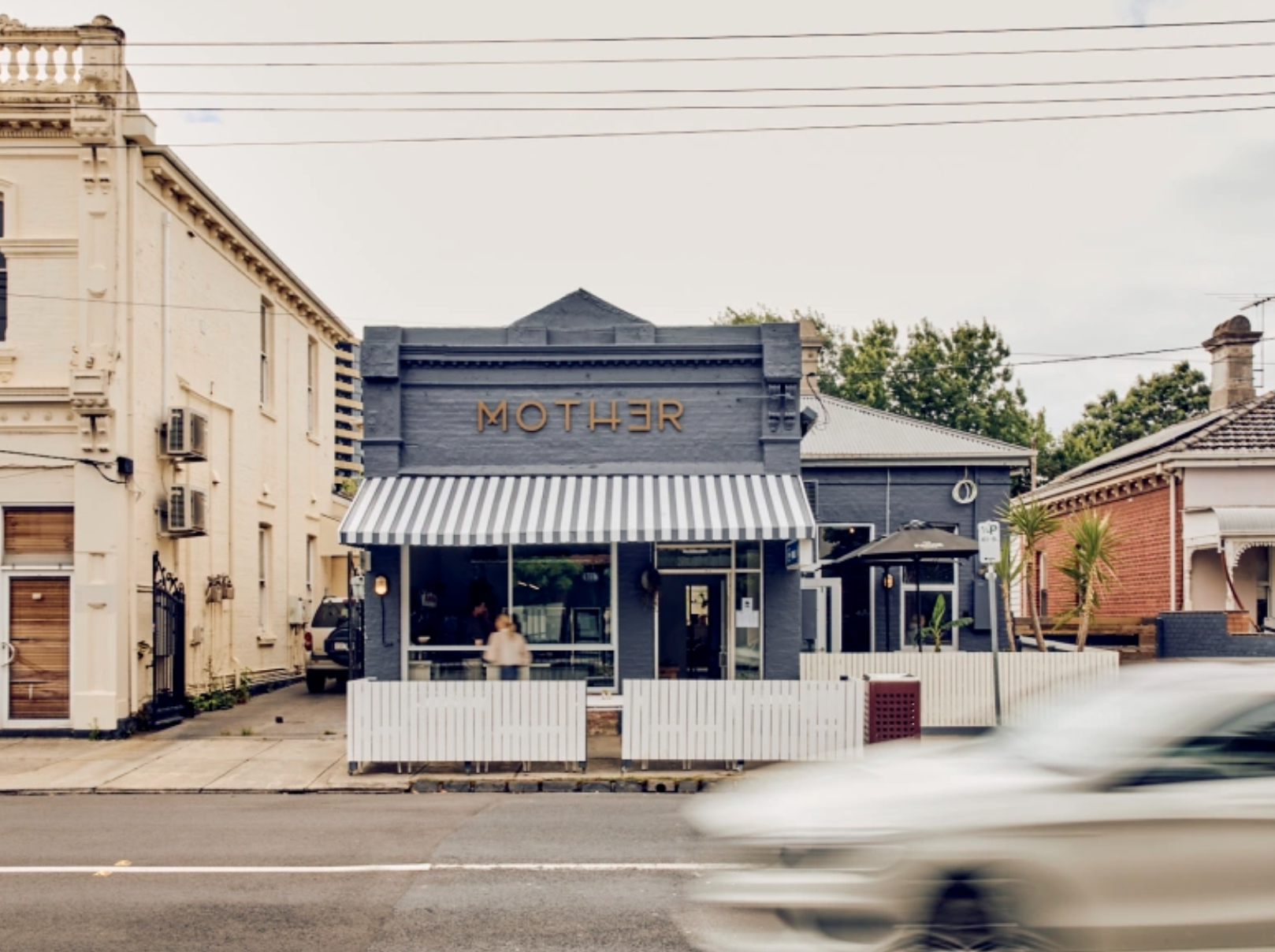 A blue cafe with the word 'MOTHER' written on it, featuring a striped awning and outdoor seating, situated on a street in Caulfield North, Melbourne with surrounding houses.