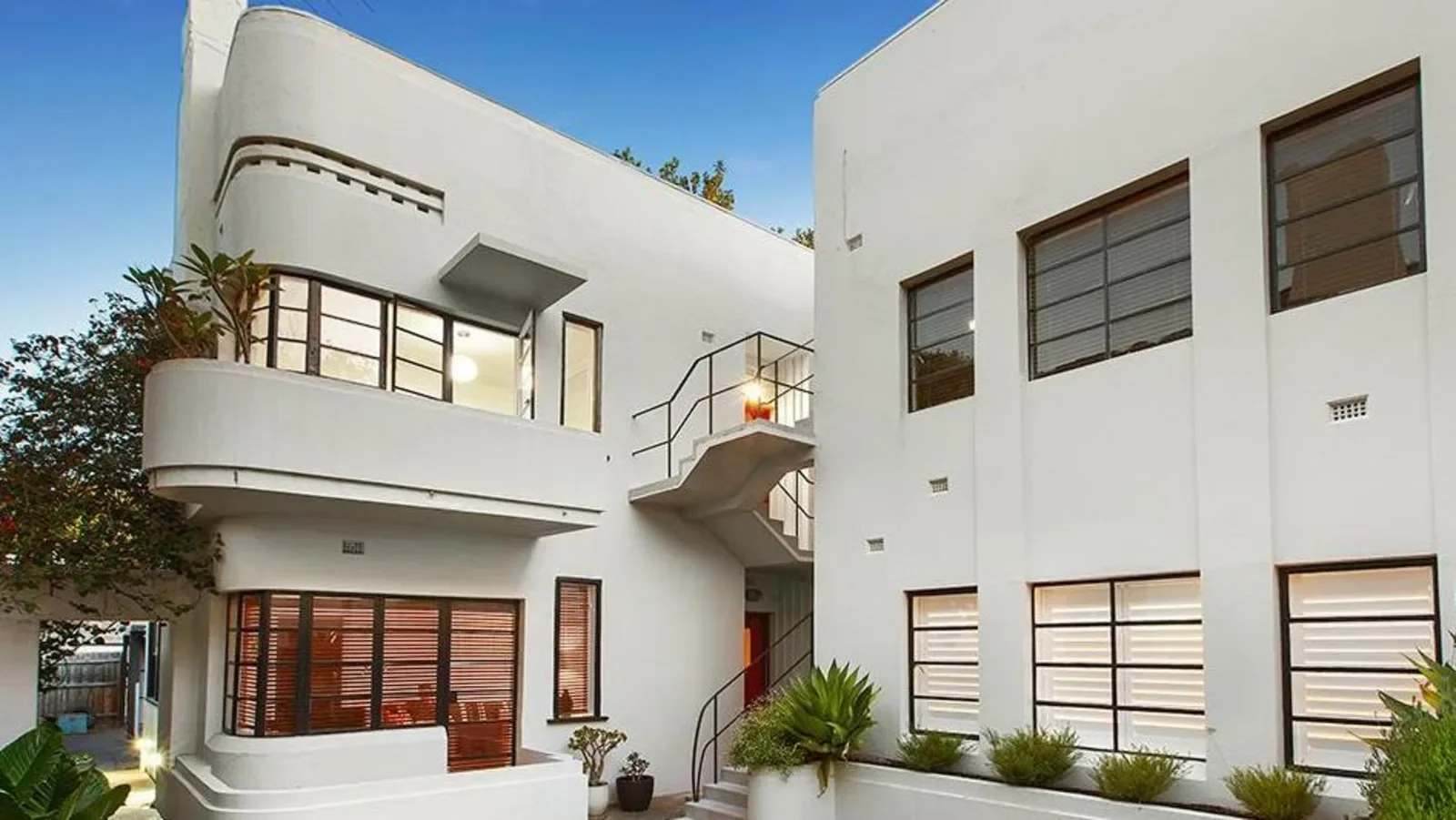 An Art Deco white apartment building in Elwood, Melbourne with large windows, outdoor staircases, and landscaped plants.