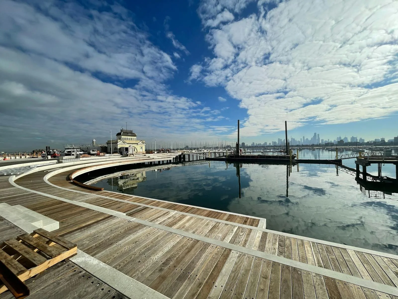 St Kilda Pier Melbourne, at the marina with a curved wooden walkway, calm water reflecting the sky and docked boats, and a city skyline in the background on a partly cloudy day.