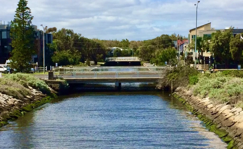 One of the Canals in Elwood, Melbourne with stone embankments on both sides, running through an urban area with buildings, trees, and bridges over the water, under a partly cloudy sky.