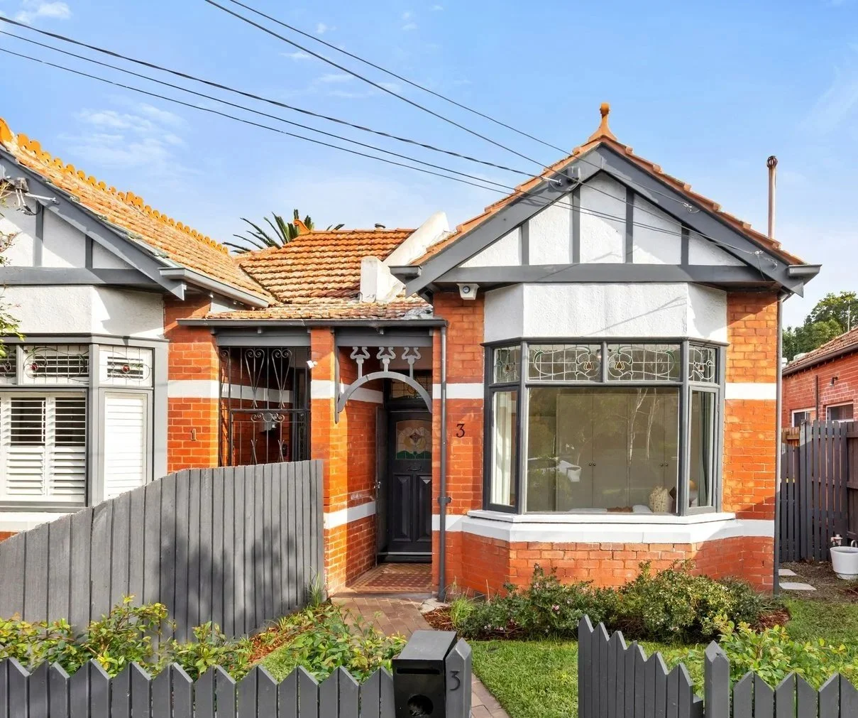 A brick house in St Kilda East, Melbourne with a gabled roof, a large front bay window, and a black front door. The house has a small front garden with plants and a gray picket fence.