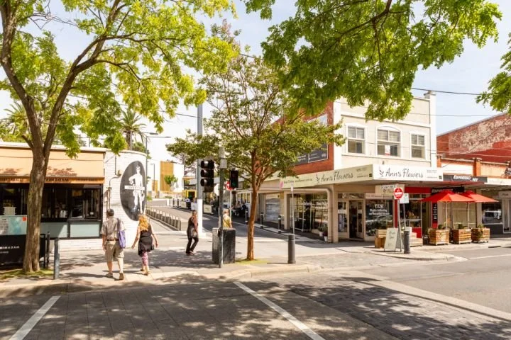 A small city street intersection in Charman Road Cheltenham, Melbourne  with four pedestrians crossing, trees shading the area, and storefronts including a flower shop and a cafe.