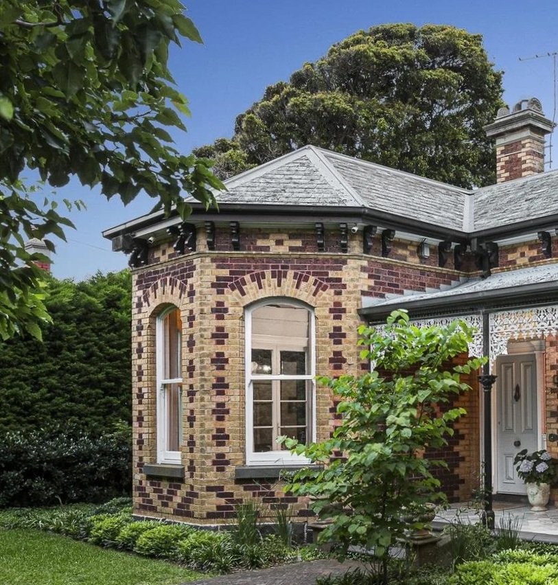 A Victorian-style brick home in Brighton, Melbourne with a rounded turret, large windows, and a well-manicured front garden with green shrubs and trees.