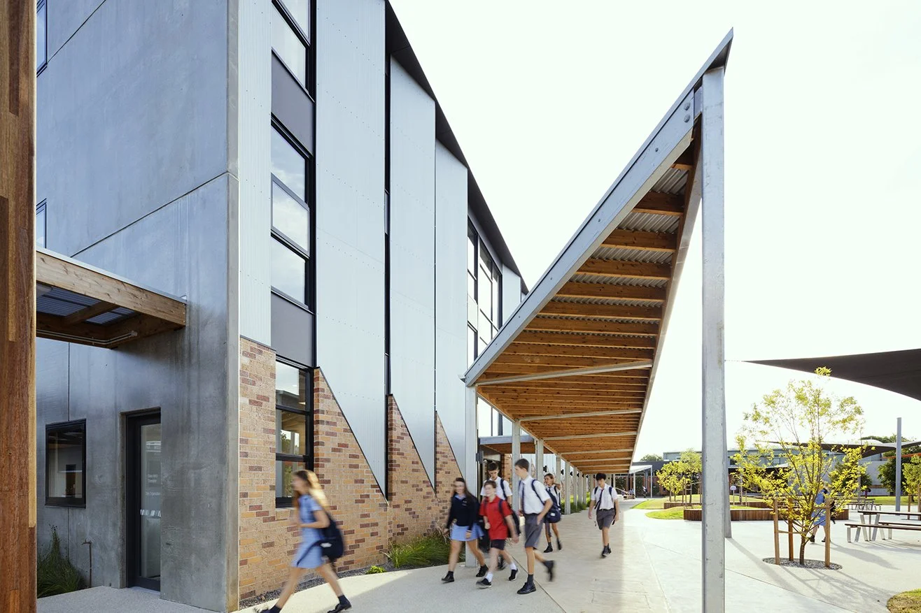 Students walking outside McKinnon High School's modern school building with brick and metal exterior, trees, and outdoor seating