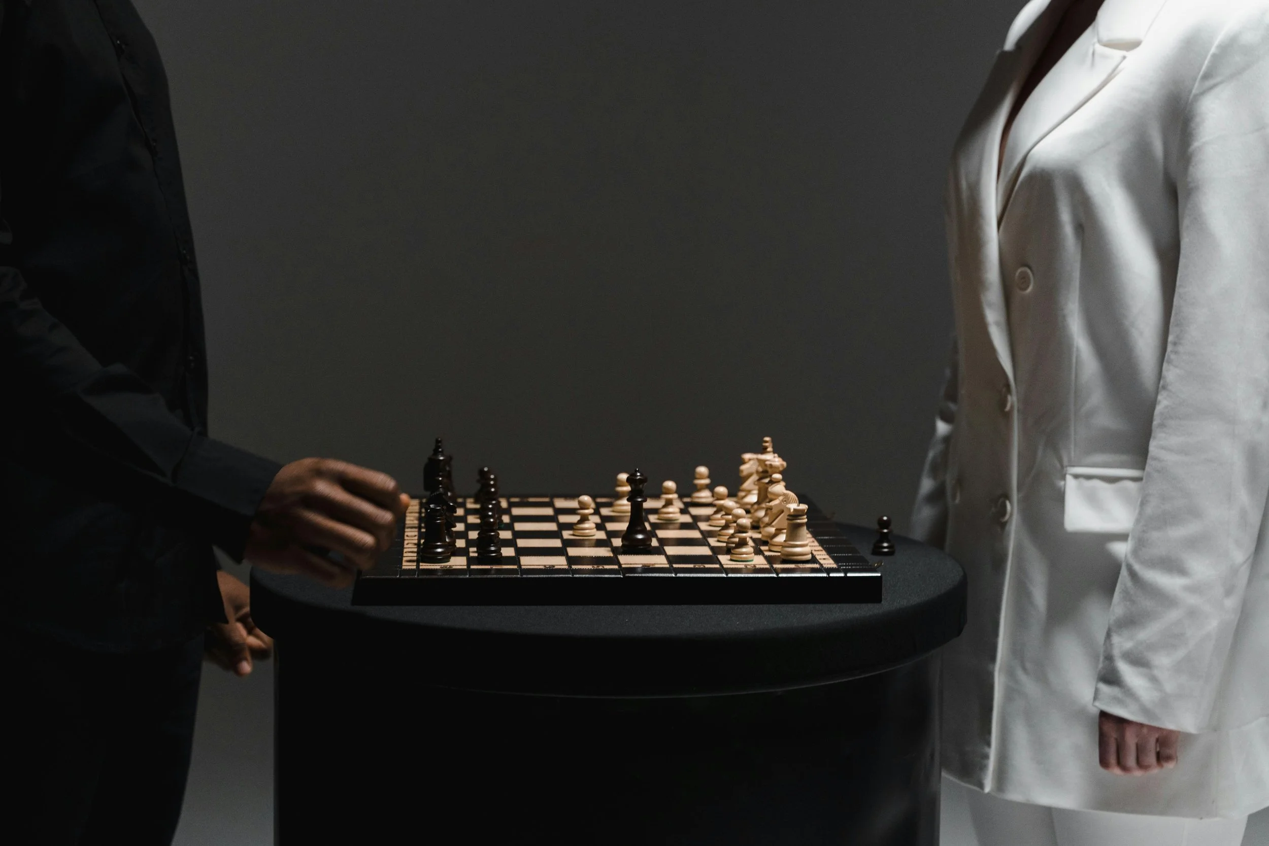 Two people playing chess on a black table, with one person in a black jacket and the other in a white blazer.