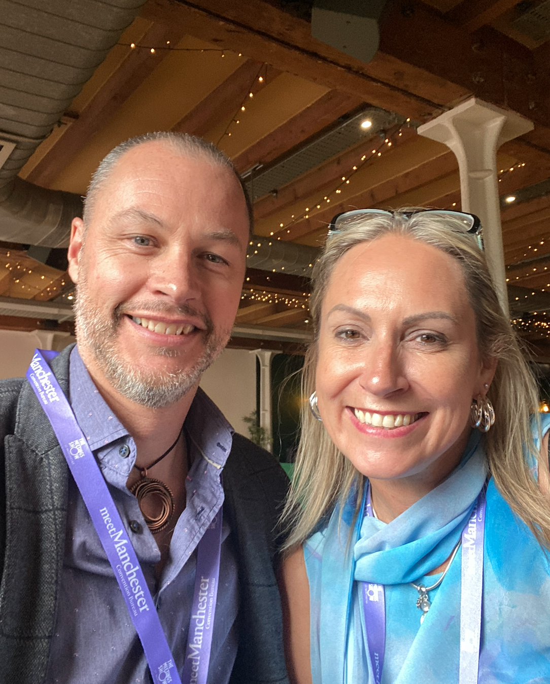A close-up photo of a smiling man and woman at an indoor event with wooden ceiling and string lights. Both are wearing conference lanyards.