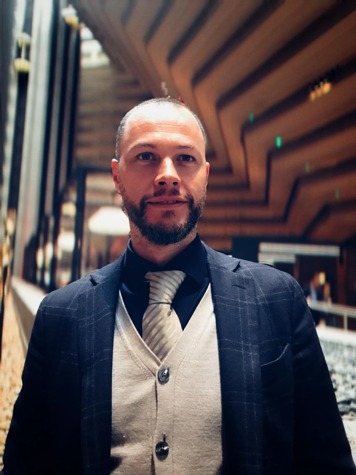 A man with a beard and short hair wearing a suit with a plaid jacket, a beige cardigan, and a striped tie, standing indoors in front of a wooden ceiling in a modern building.