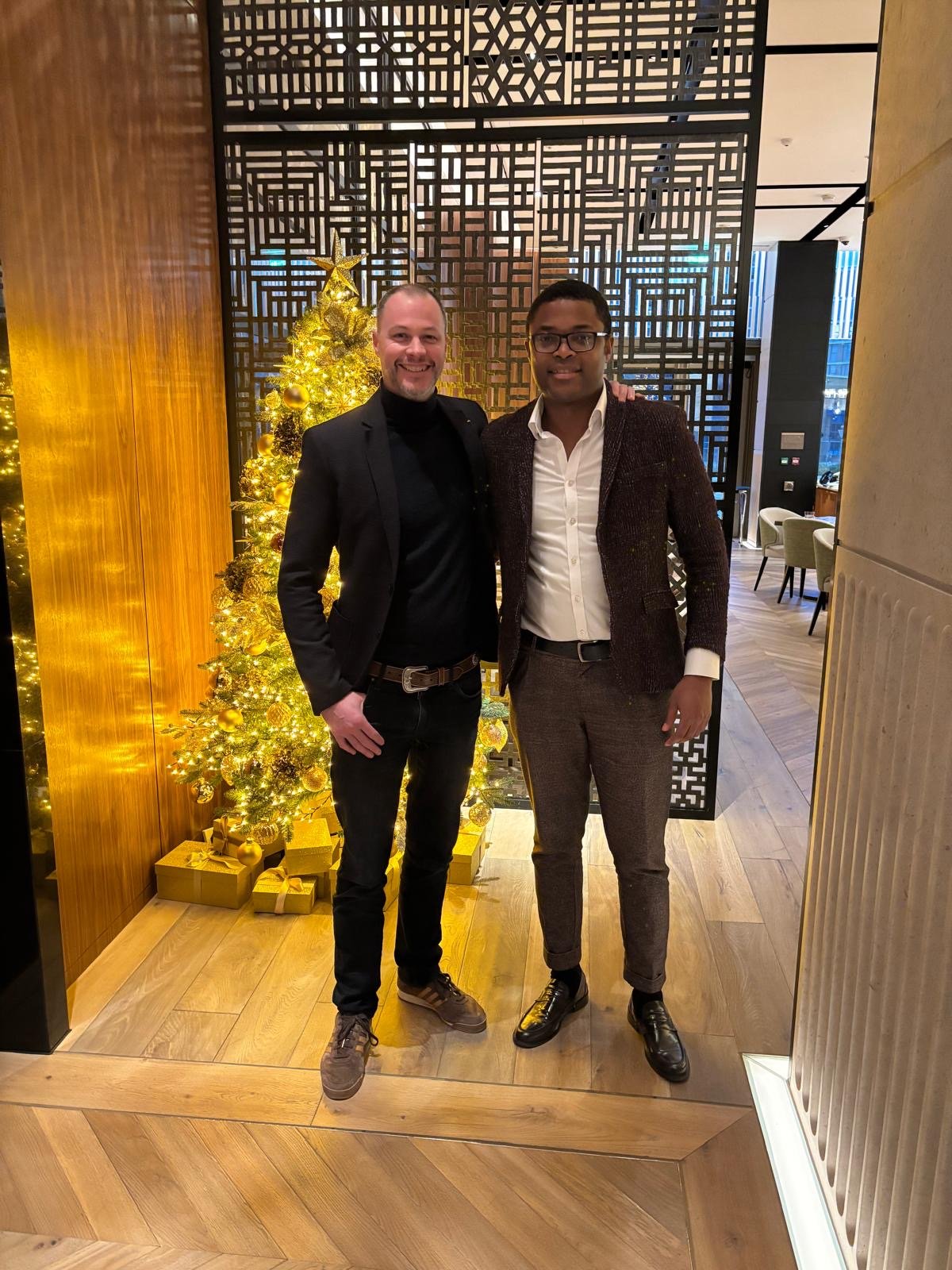 Two men in formal attire standing indoors near a decorated Christmas tree with golden ornaments and wrapped gifts, with a wooden and geometric patterned background.