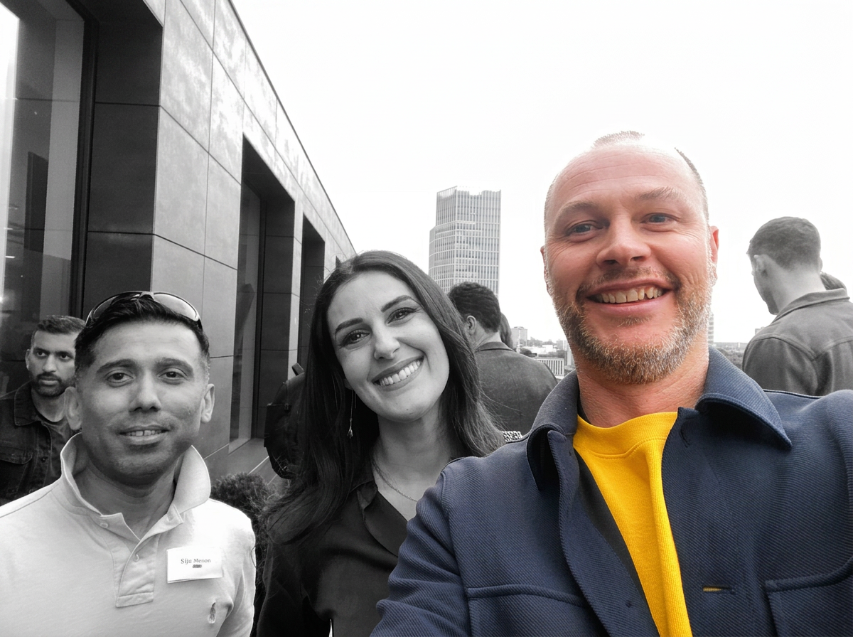 Three people smiling and taking a selfie on a rooftop with a city skyline in the background. The photo is mostly black and white, with the exception of the man's yellow shirt and the woman's earrings.