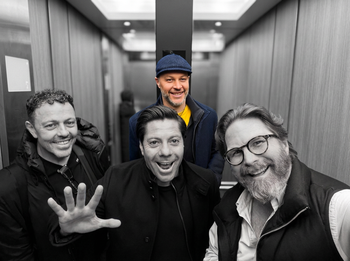 Four men smiling and looking at the camera inside an elevator, with one man in the front making a peace sign. The photo is mostly black and white, except for one man wearing a yellow shirt and blue hat, who is in color.