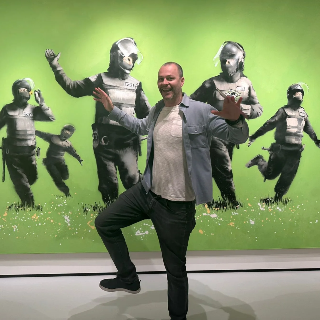 A man standing in front of a mural with police officers in riot gear. He is smiling and making a playful pose with his hands.