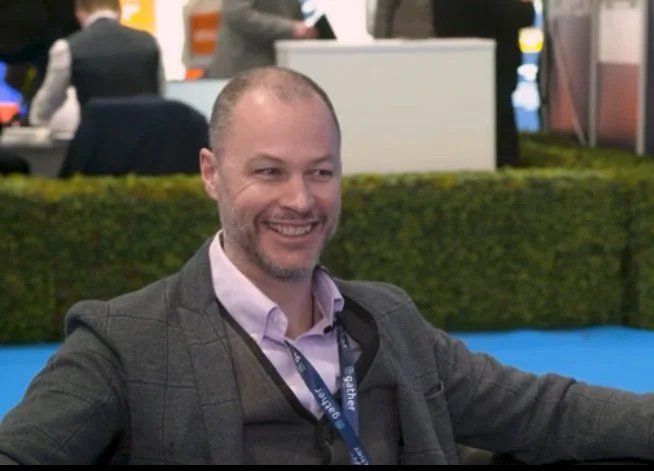 Smiling man in business attire sitting at a conference or event, with other people and booths in the background.