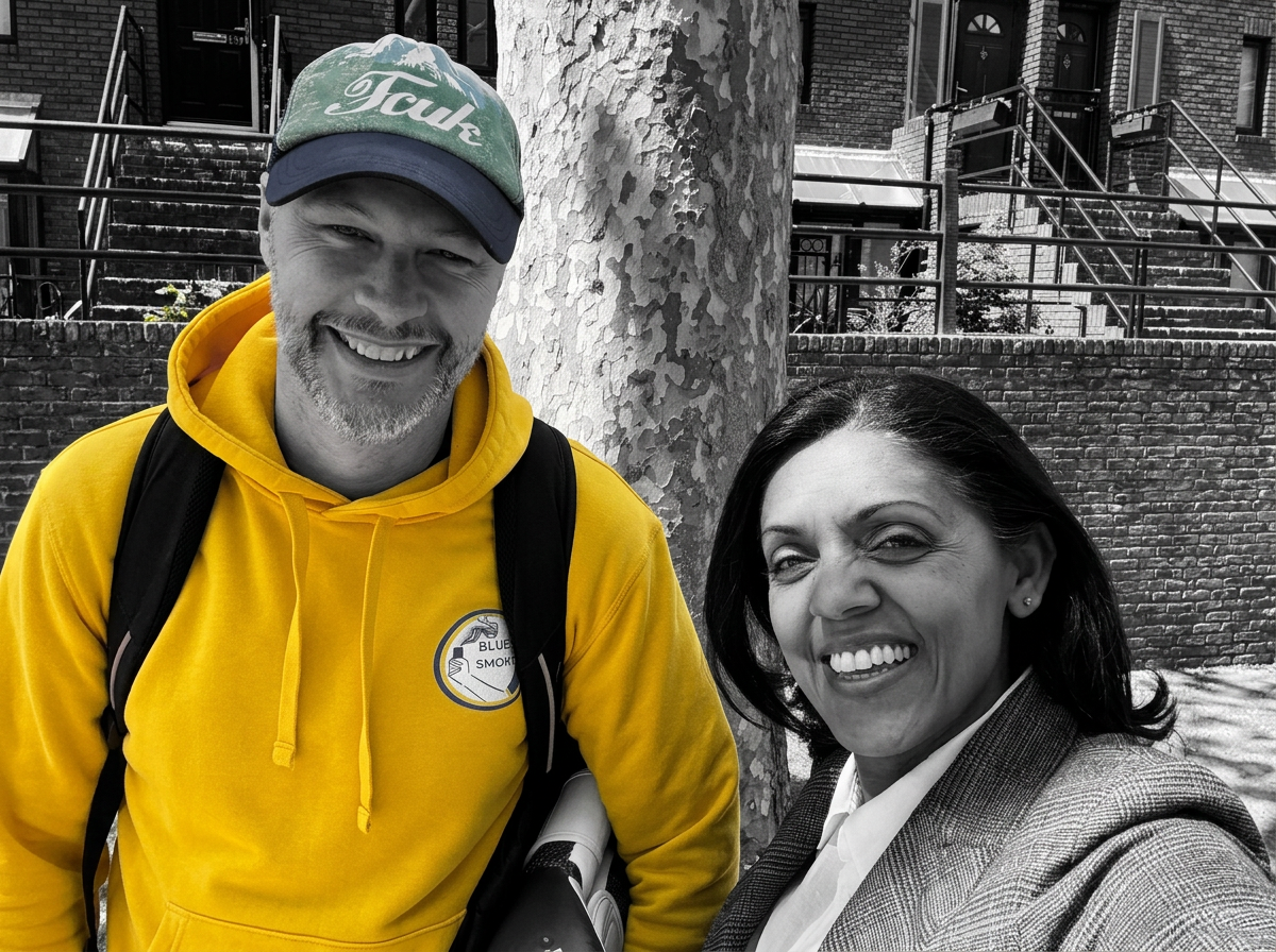 A smiling man in a yellow hoodie and a woman in a blazer are outdoors, standing next to a tree, with buildings and stairs in the background.